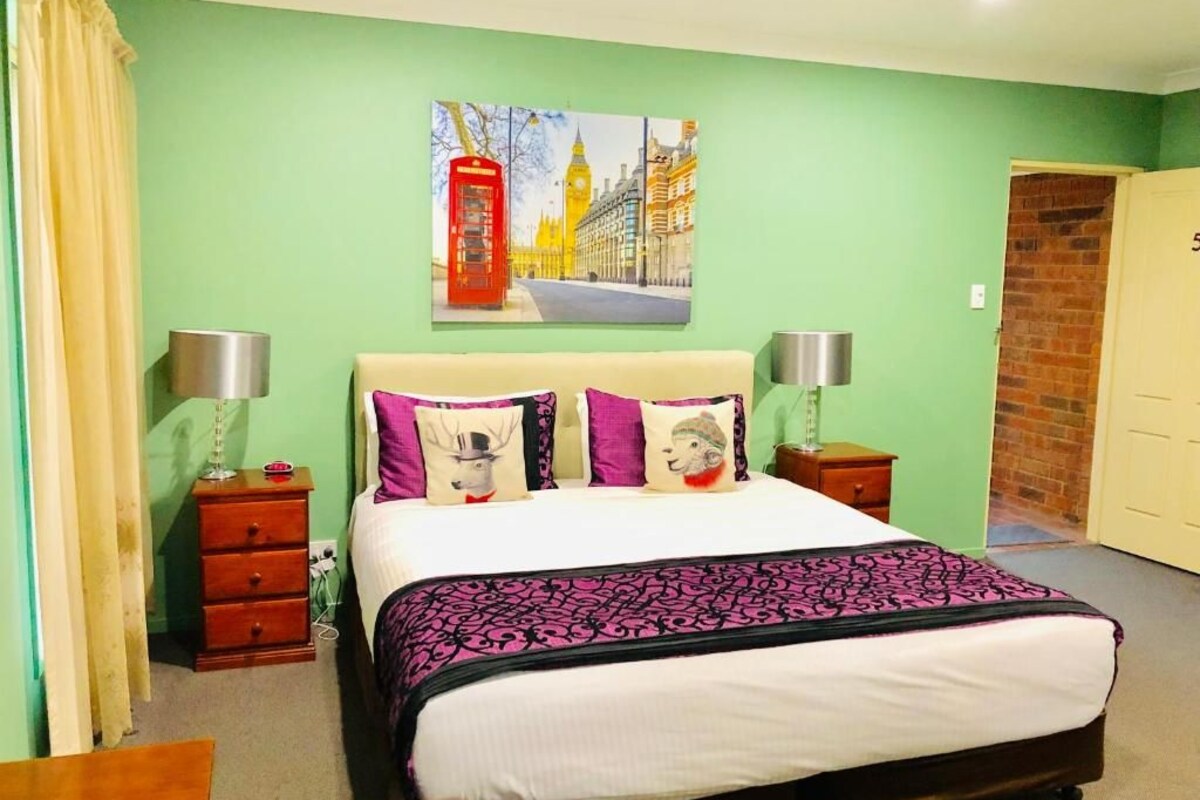 A bedroom with a bed, two bedside tables, a lamp, a painting of London landmarks, and a brick wall.