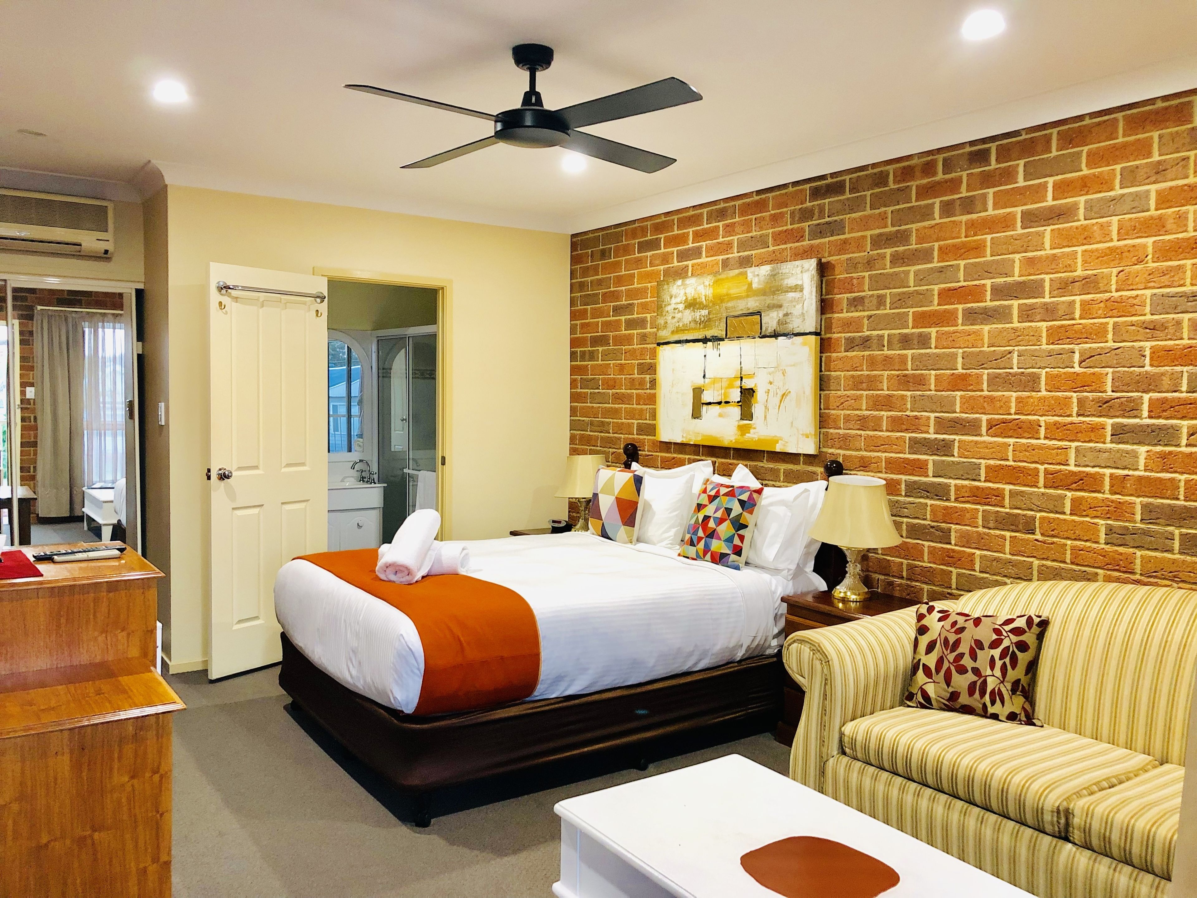 A hotel room with a brick wall, a bed with white bedding and orange accents, a wooden desk, a striped sofa, and a ceiling fan.