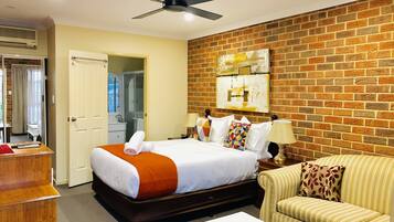 A hotel room with a brick wall, a bed with white bedding and orange accents, a wooden desk, a striped sofa, and a ceiling fan.