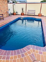 Una piscina rectangular con interior azul, rodeada por un patio de ladrillo y plantas en macetas.