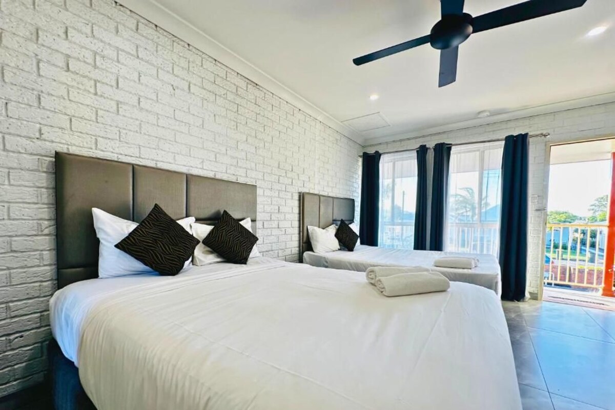A hotel room with a large bed, a ceiling fan, a brick wall, and a view of the outdoors.