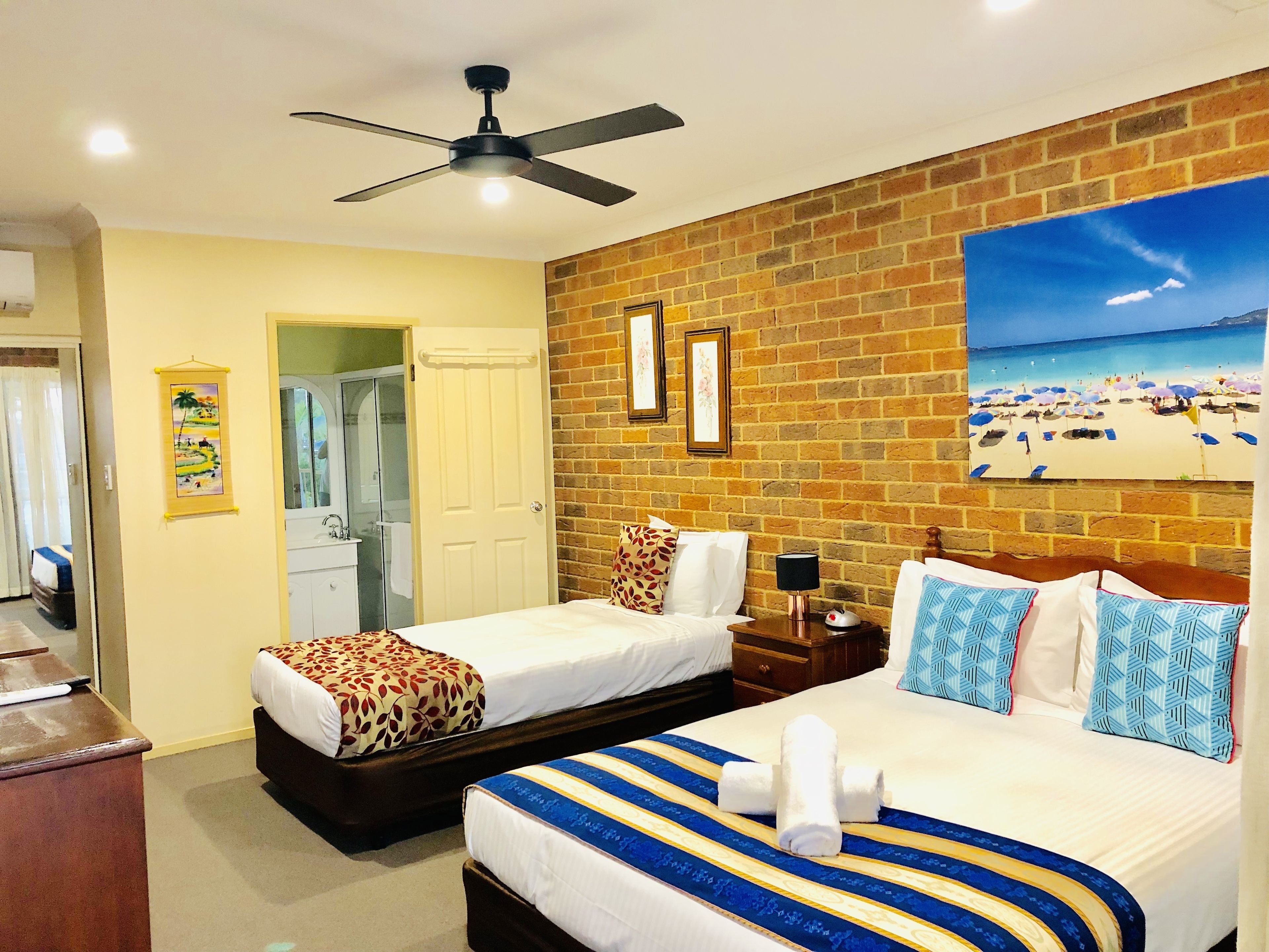 A hotel room with two beds, a ceiling fan, a bathroom with a mirror, and a beach view painting on the wall.