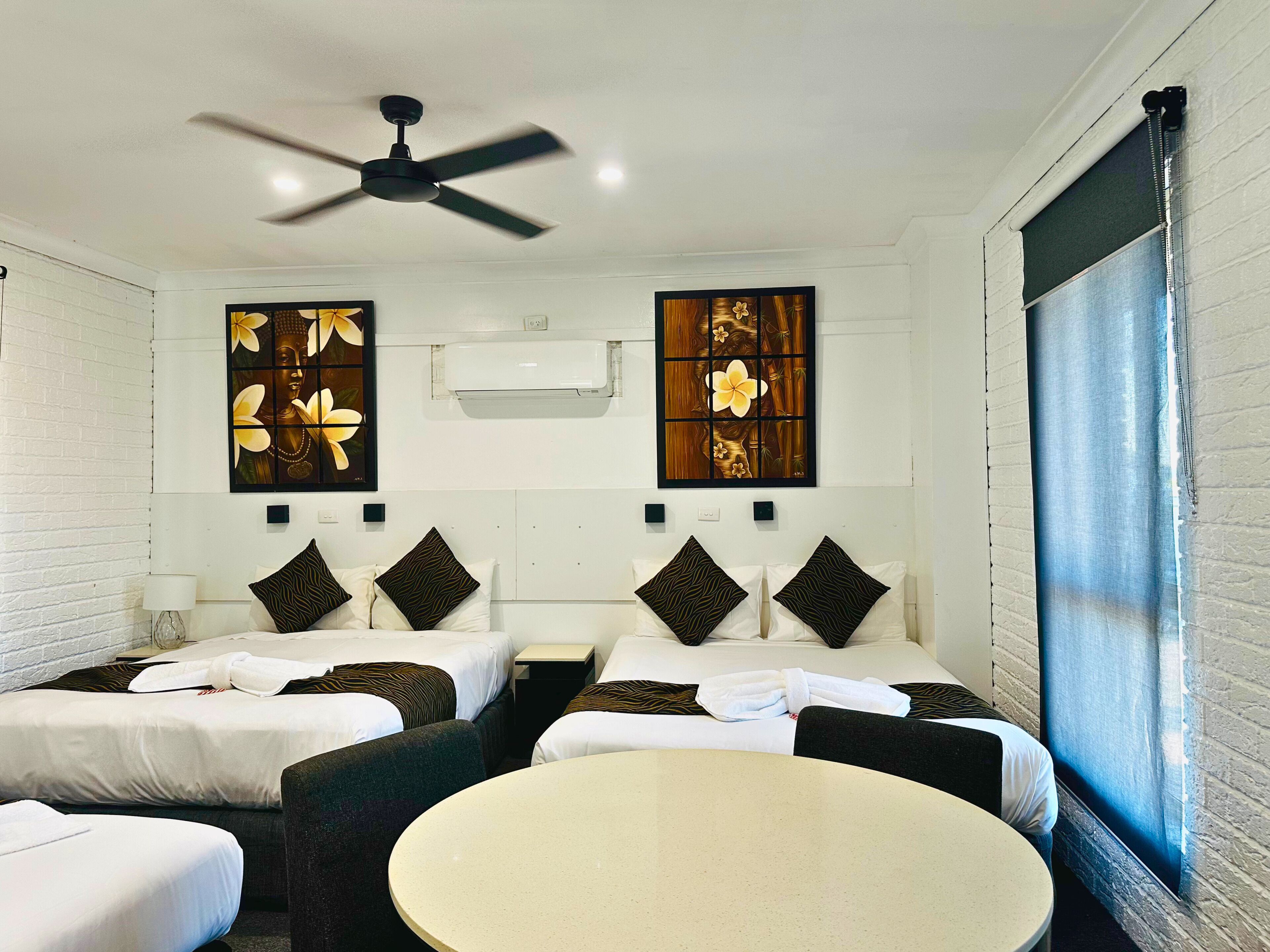A hotel room with two beds, a round table, a ceiling fan, and artwork on the walls.