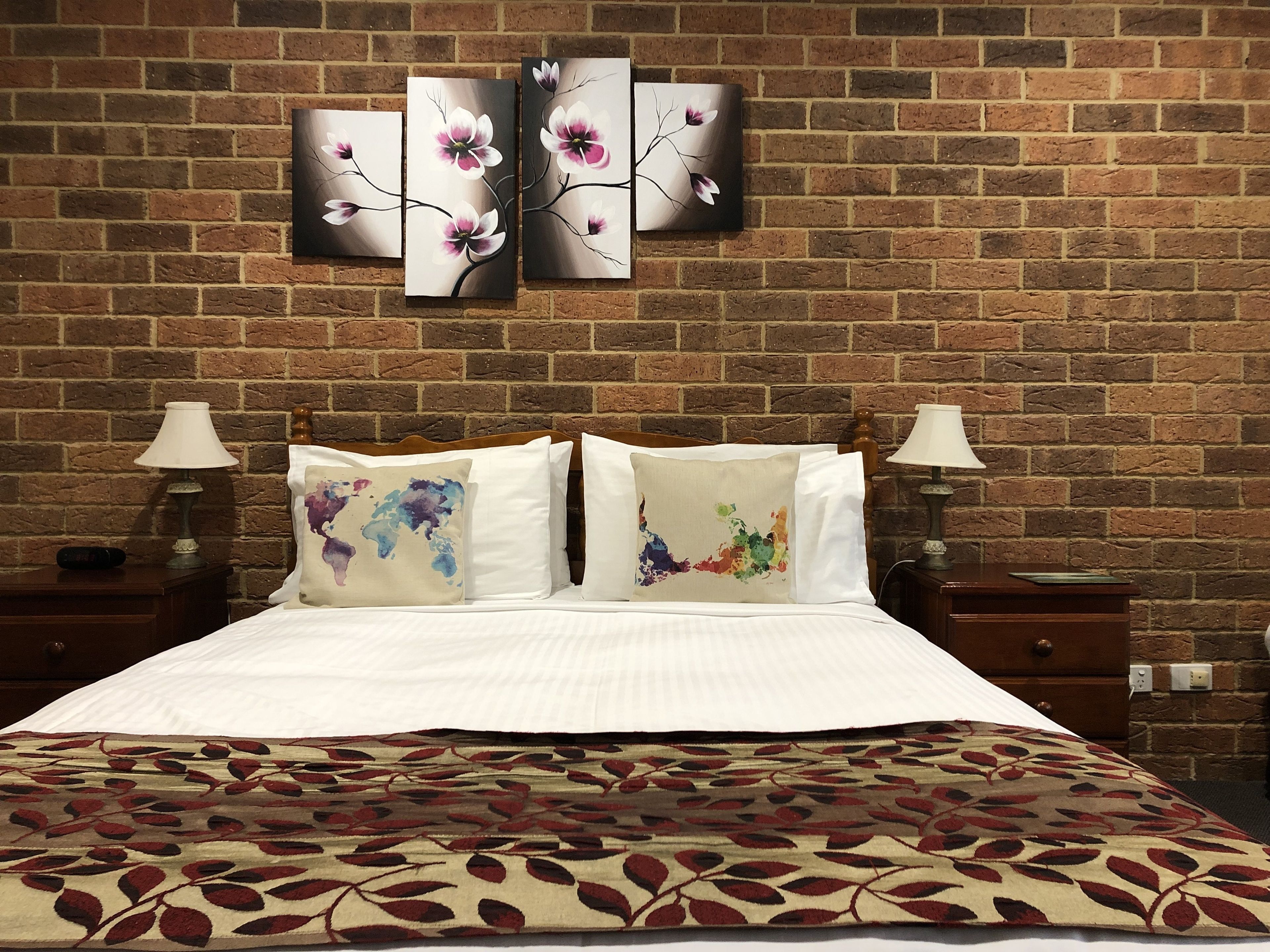 A hotel room with a brick wall, a bed with a floral bedspread, two nightstands with lamps, and three wall-mounted paintings.