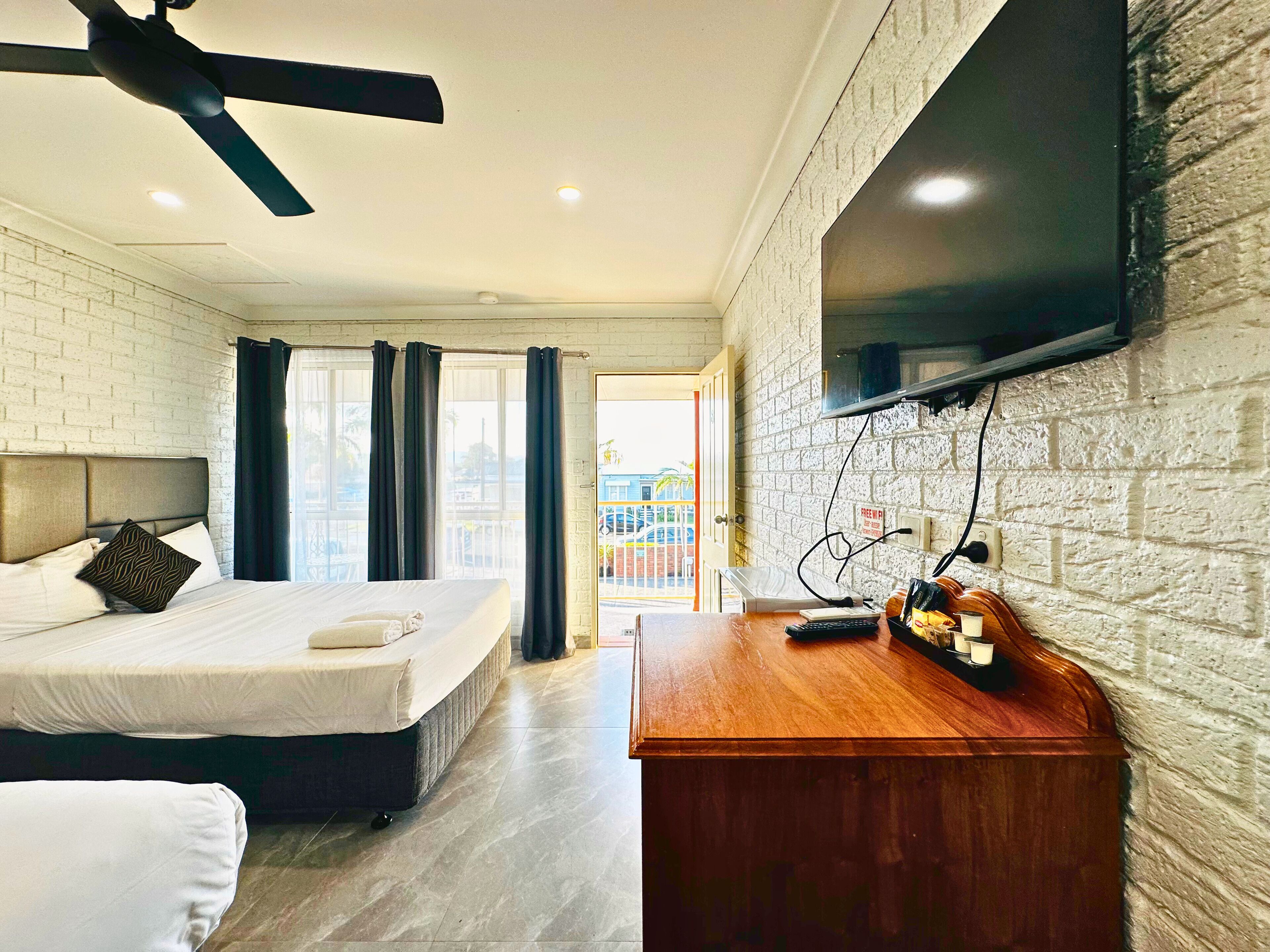 Habitación de hotel con cama, escritorio con televisor de pantalla plana, ventilador de techo y pared de ladrillo.