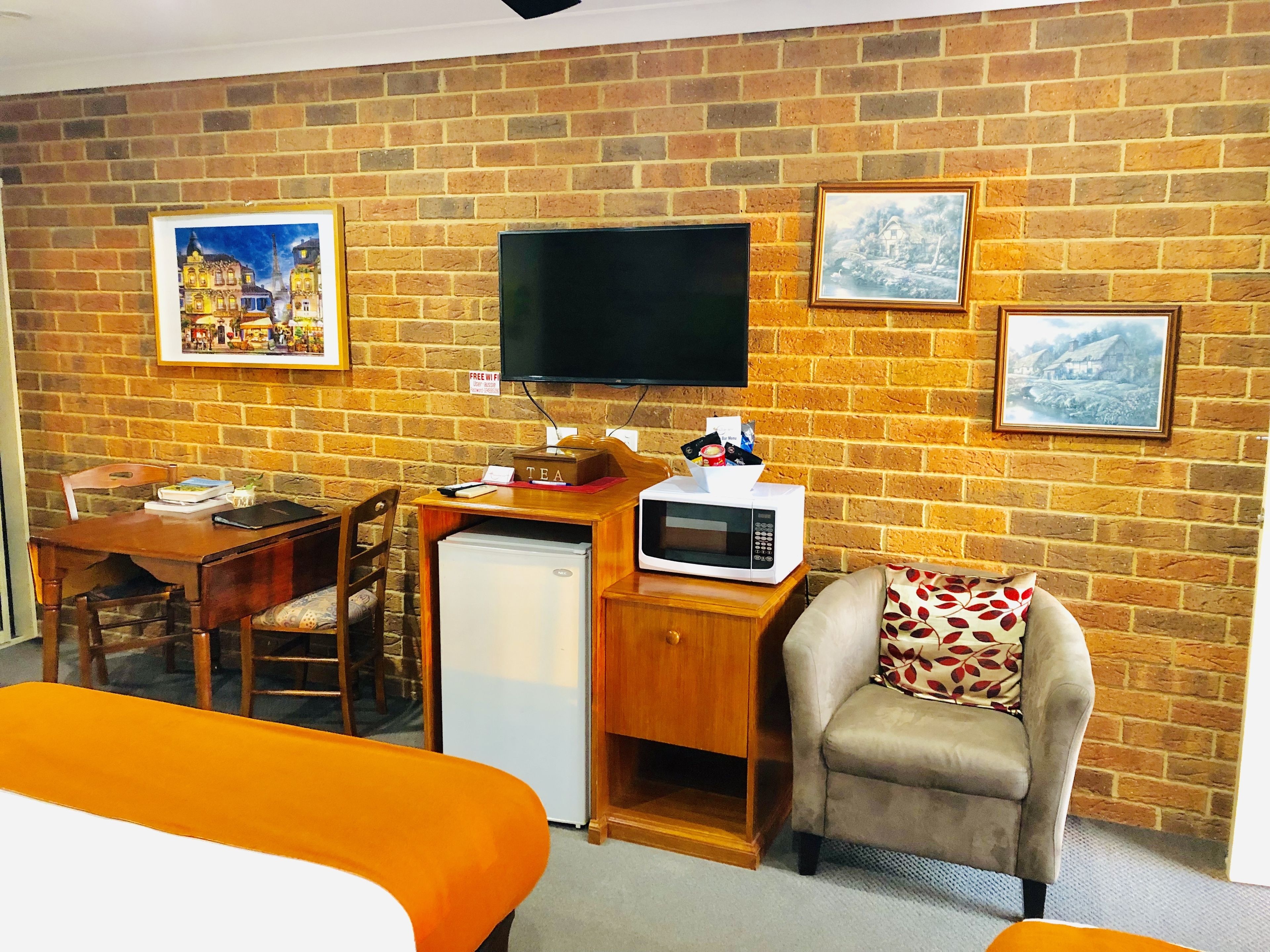 A room with a brick wall, a flat-screen TV, a small refrigerator, a microwave, a desk with a chair, and a sofa with a patterned pillow.
