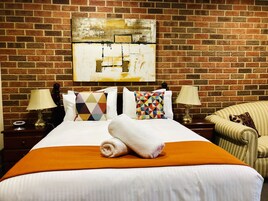 Quarto de hotel com parede de tijolos, cama com roupa de cama branca, duas mesas de cabeceira com luminárias e uma poltrona listrada.