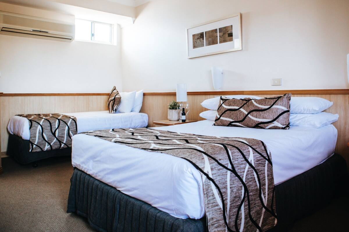 Une chambre d’hôtel avec deux lits, une fenêtre, un appareil de climatisation, une table de chevet avec une lampe et un tableau encadré au mur.