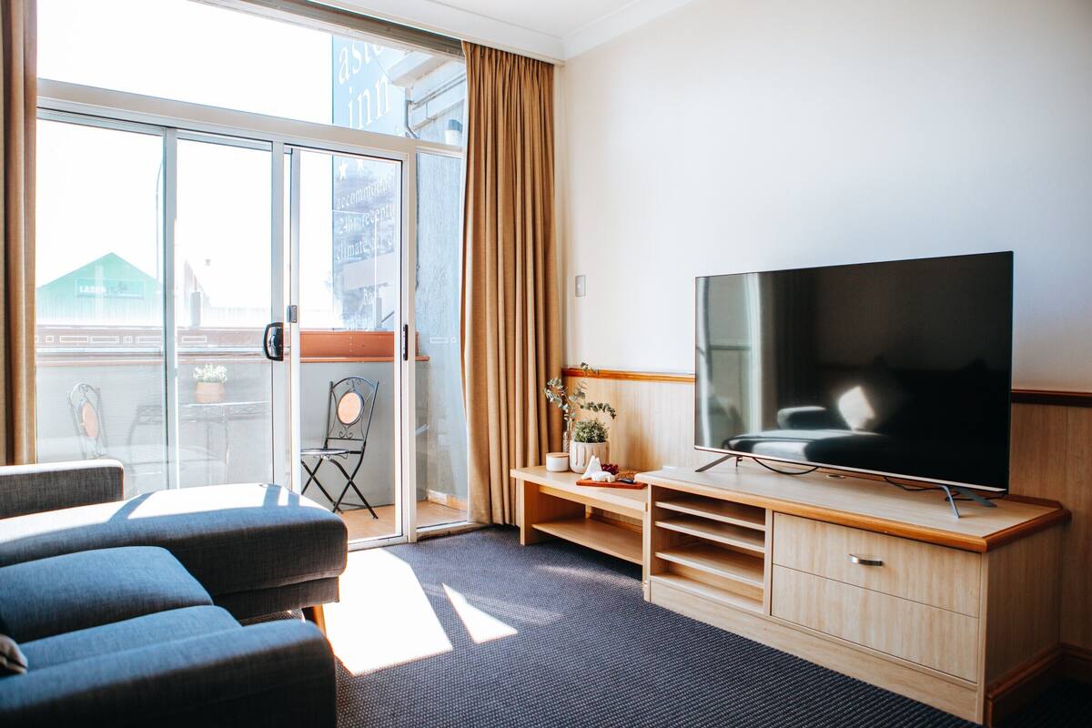 Une chambre d’hôtel moderne dotée d’un grand téléviseur à écran plat, d’un canapé, d’un meuble de rangement en bois et d’une porte-fenêtre coulissante donnant sur un balcon.