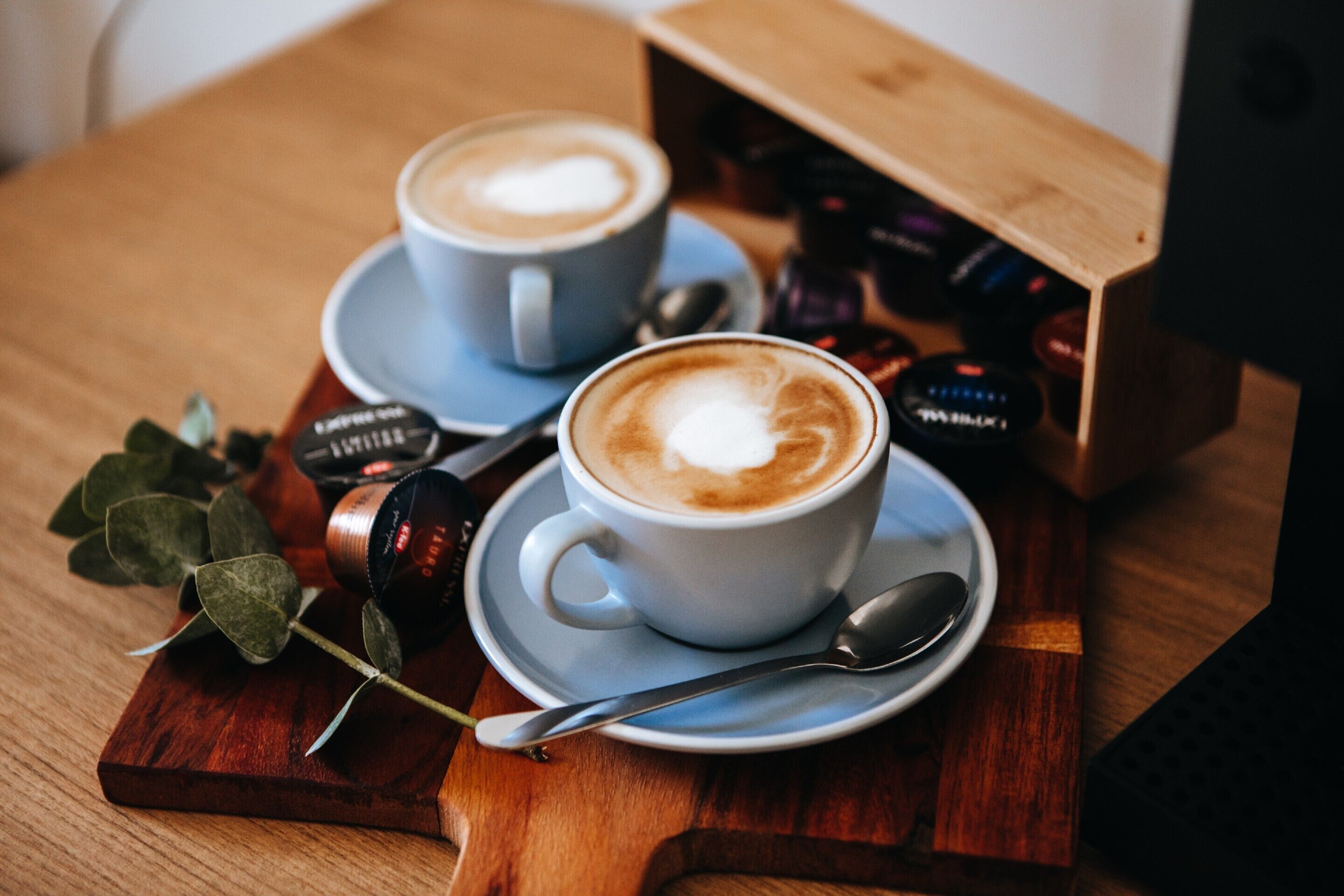 Deux tasses de café sur un plateau en bois, accompagnées d’une cuillère et d’une petite plante.