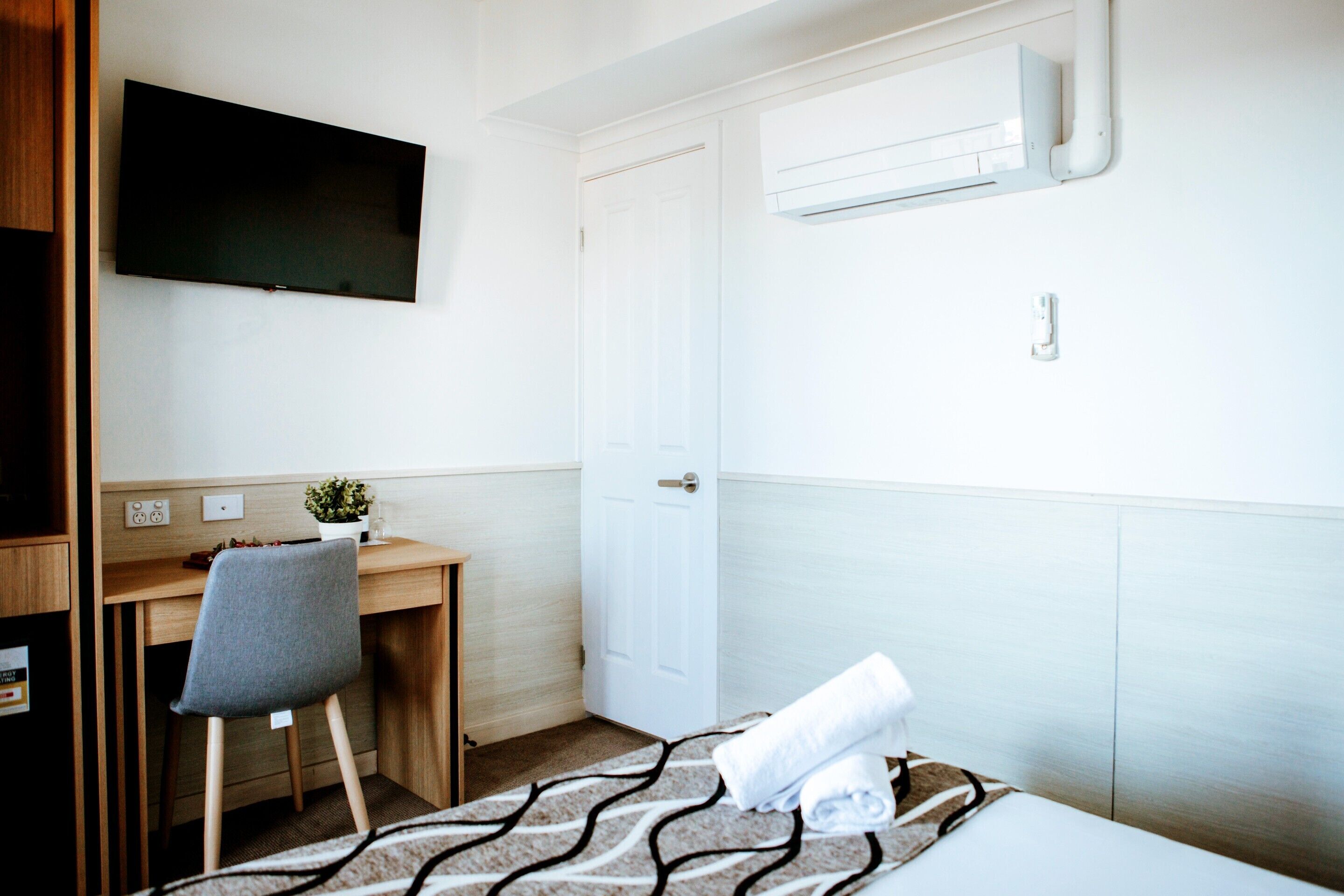 A hotel room with a bed, a desk with a chair, a television mounted on the wall, and a door.