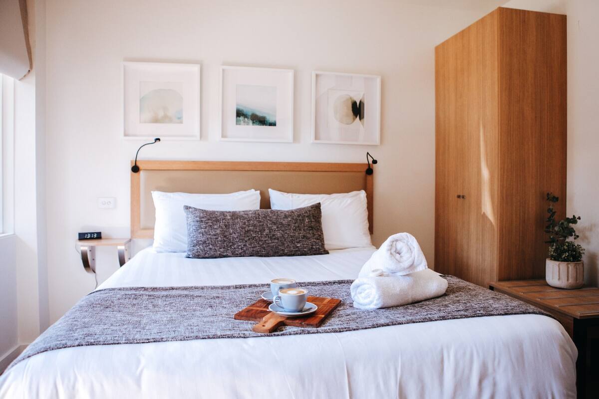 Un lit bien fait, avec des draps blancs, deux oreillers, une couverture et un plateau sur lequel reposent deux tasses de café. Trois tableaux encadrés sont accrochés au mur, au-dessus du lit.