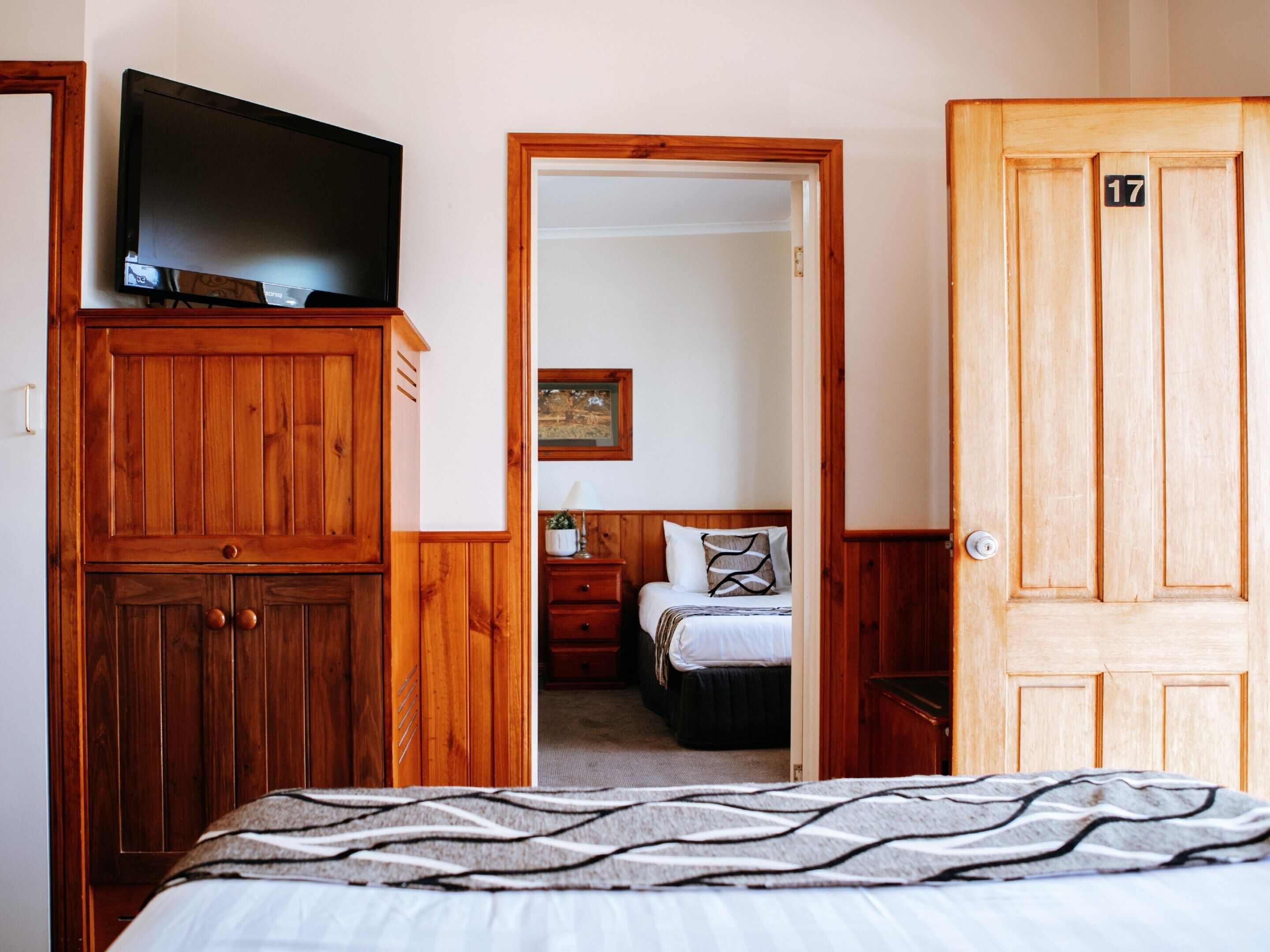 A bedroom with a bed, a television on a wooden cabinet, a wooden door with the number 17, and a hallway leading to another room.