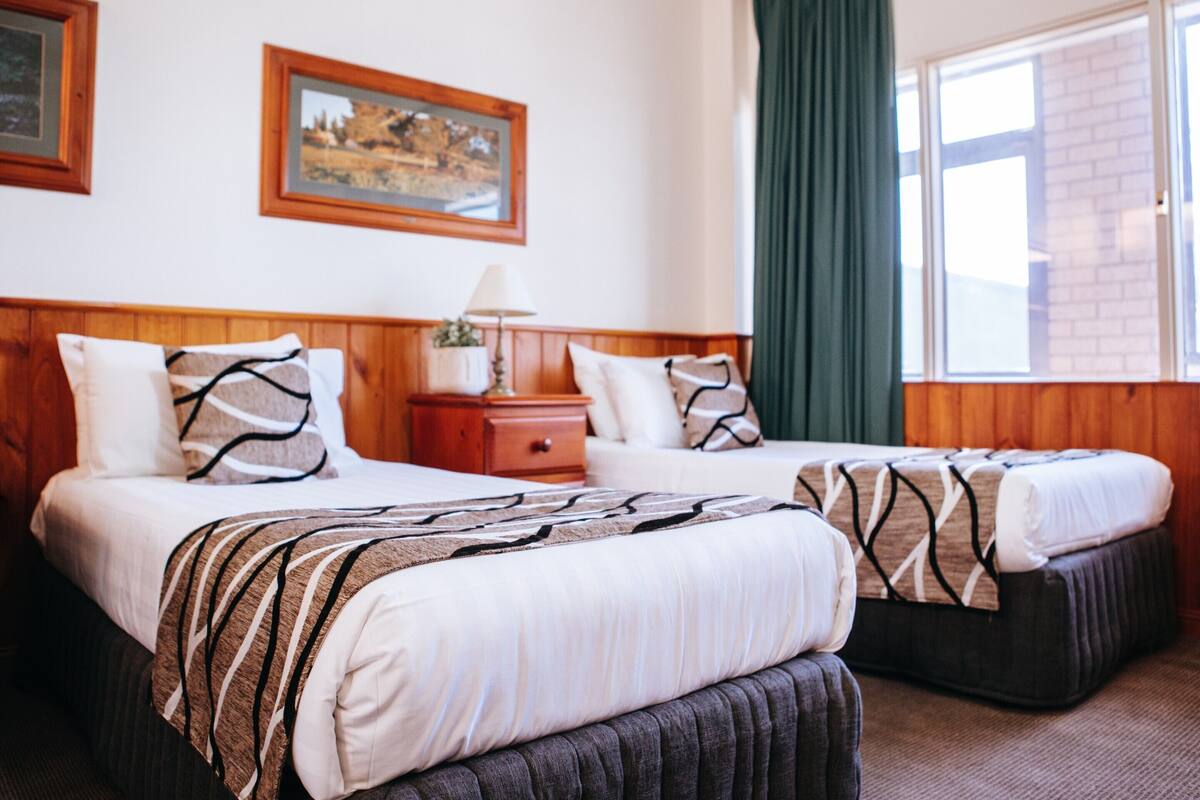 Une chambre d’hôtel avec deux lits, une table de chevet, une lampe, un tableau au mur et une fenêtre avec des rideaux.