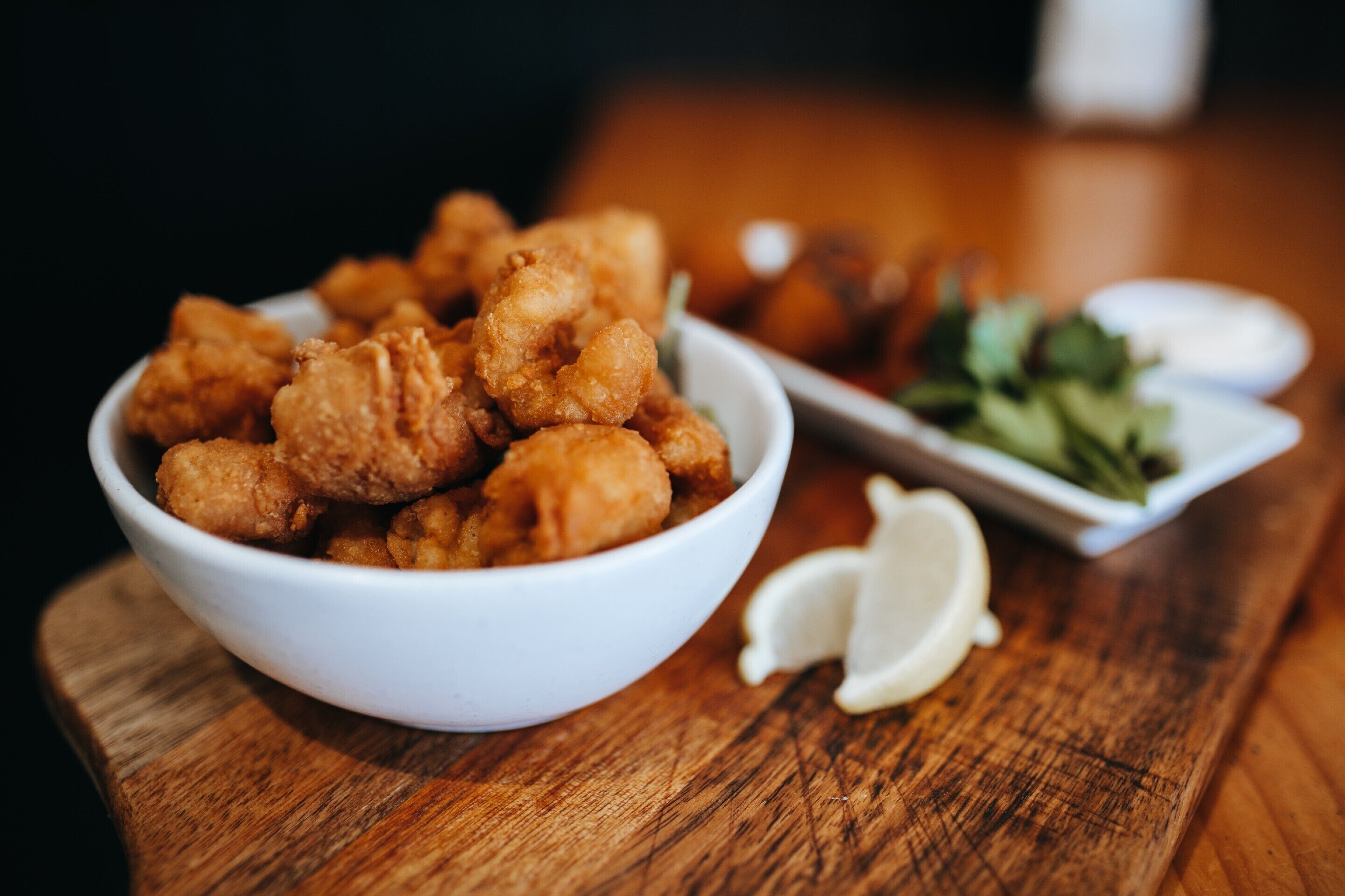 A bowl of fried chicken on a wooden table with a small plate of greens and lemon wedges.