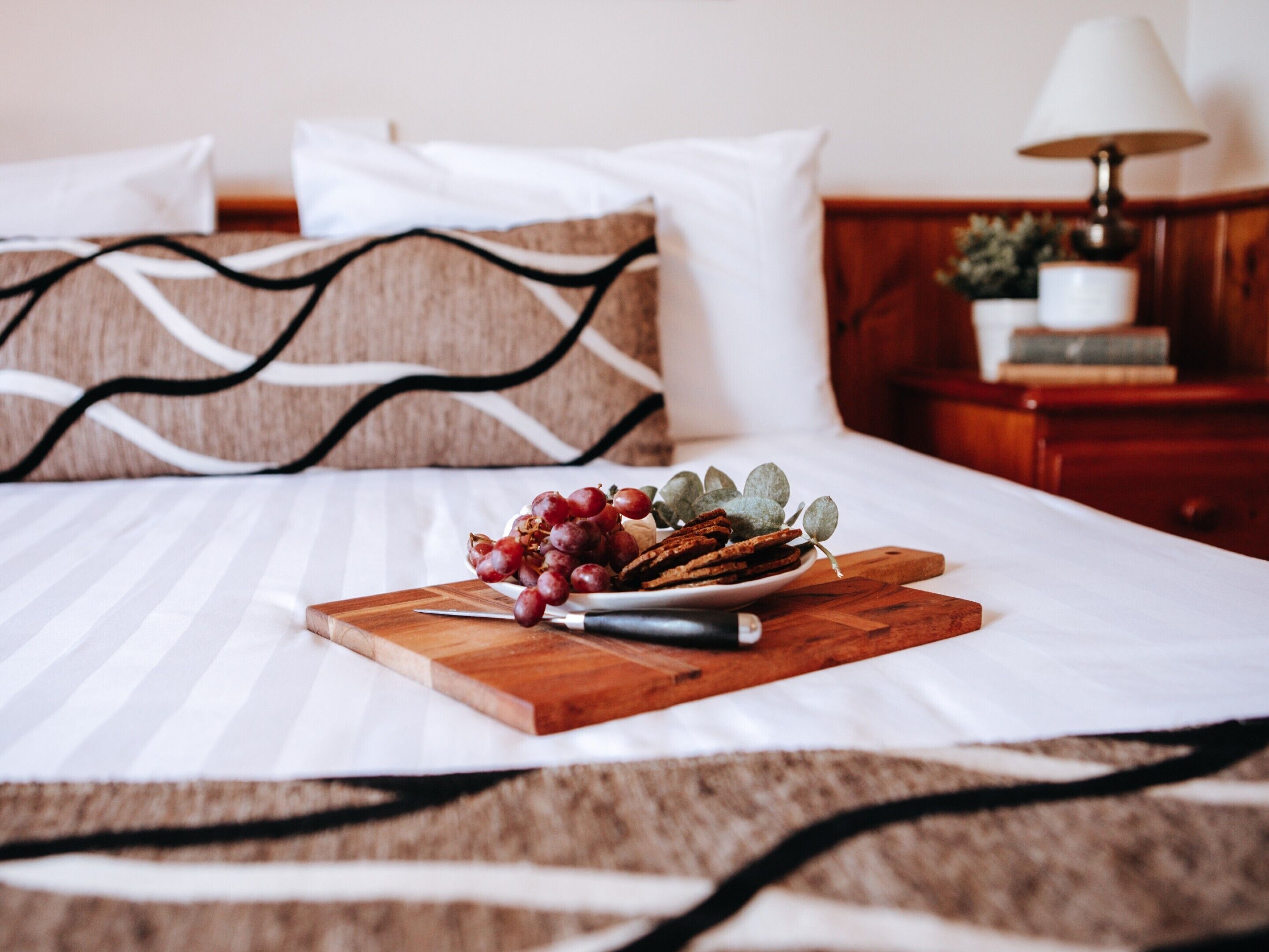 A neatly made bed with a wooden cutting board holding grapes and crackers, a lamp on a bedside table, and a plant in a white pot.