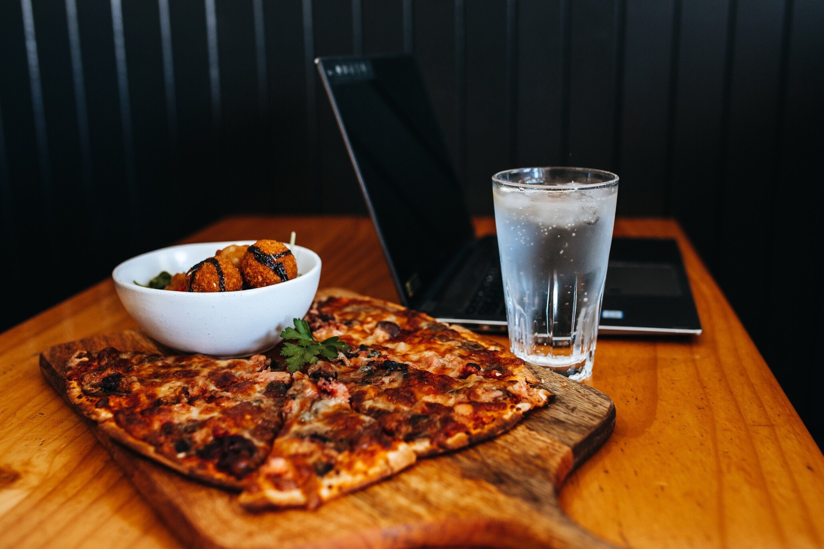 En pizza på en träbricka, ett glas vatten och en skål med mat på ett bord.