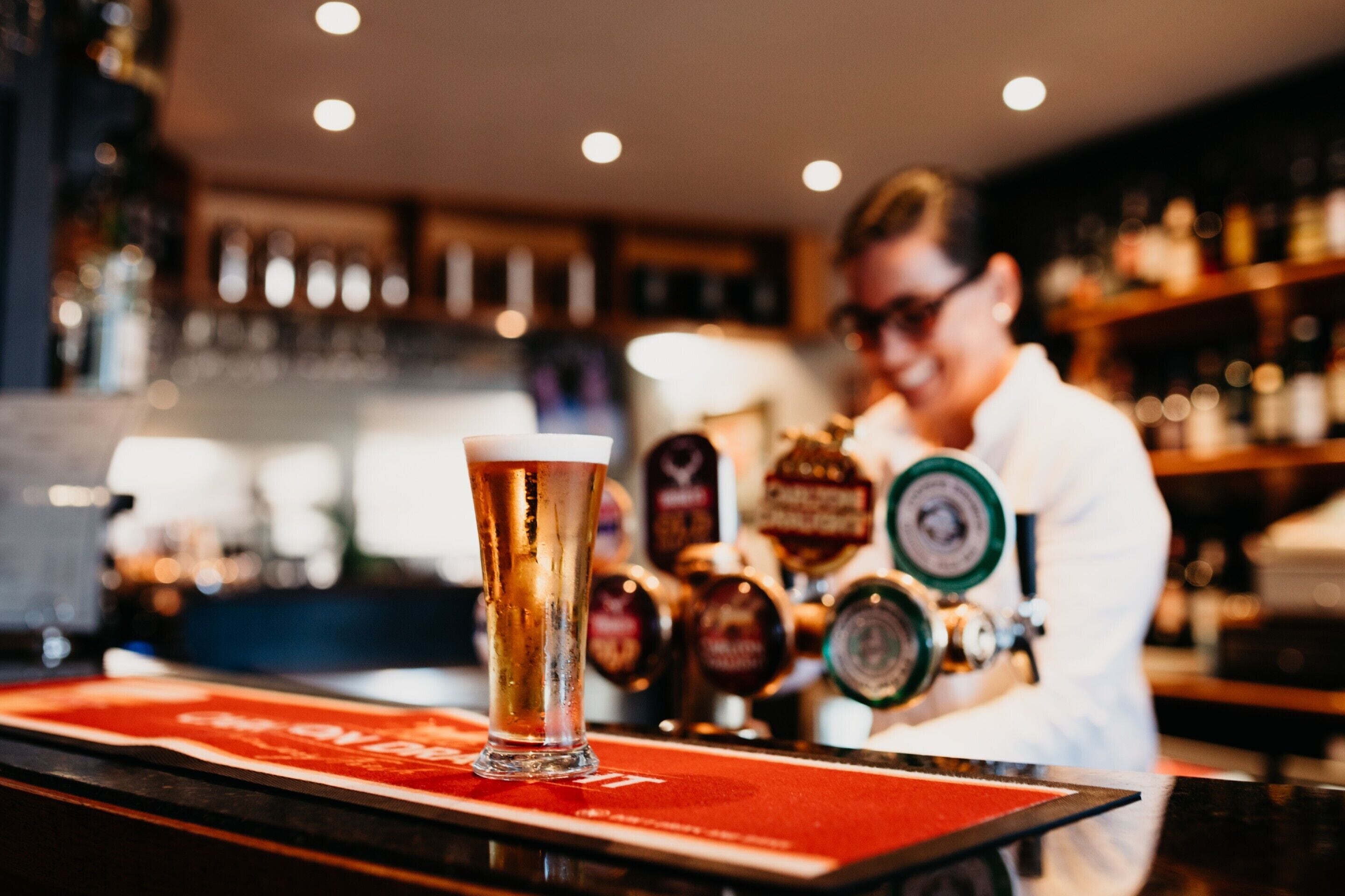 Eine Bar mit einem hohen Glas Bier auf einem roten Untersetzer, ein Barkeeper im Hintergrund und verschiedene Zapfhähne.