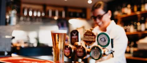 A bar with a tall glass of beer on a red coaster, a bartender in the background, and various beer taps.