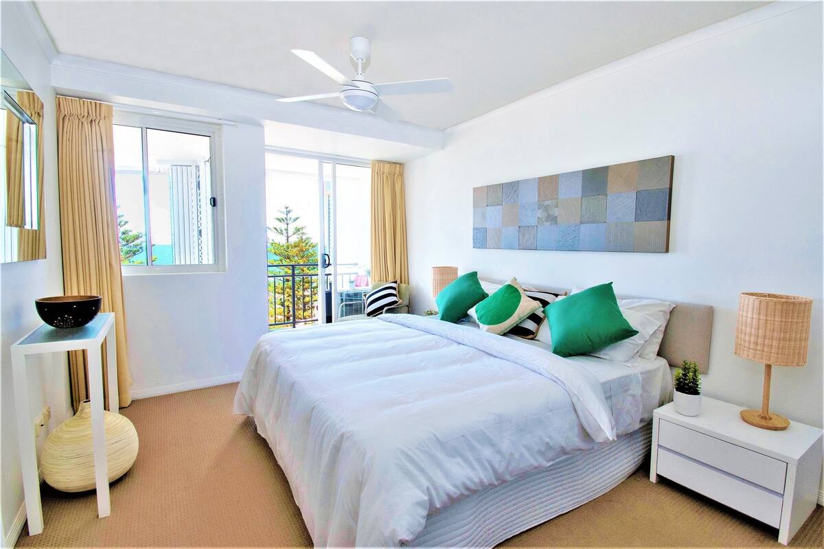 Quarto bem arrumado com cama, mesa de cabeceira, abajur e vista para o exterior.