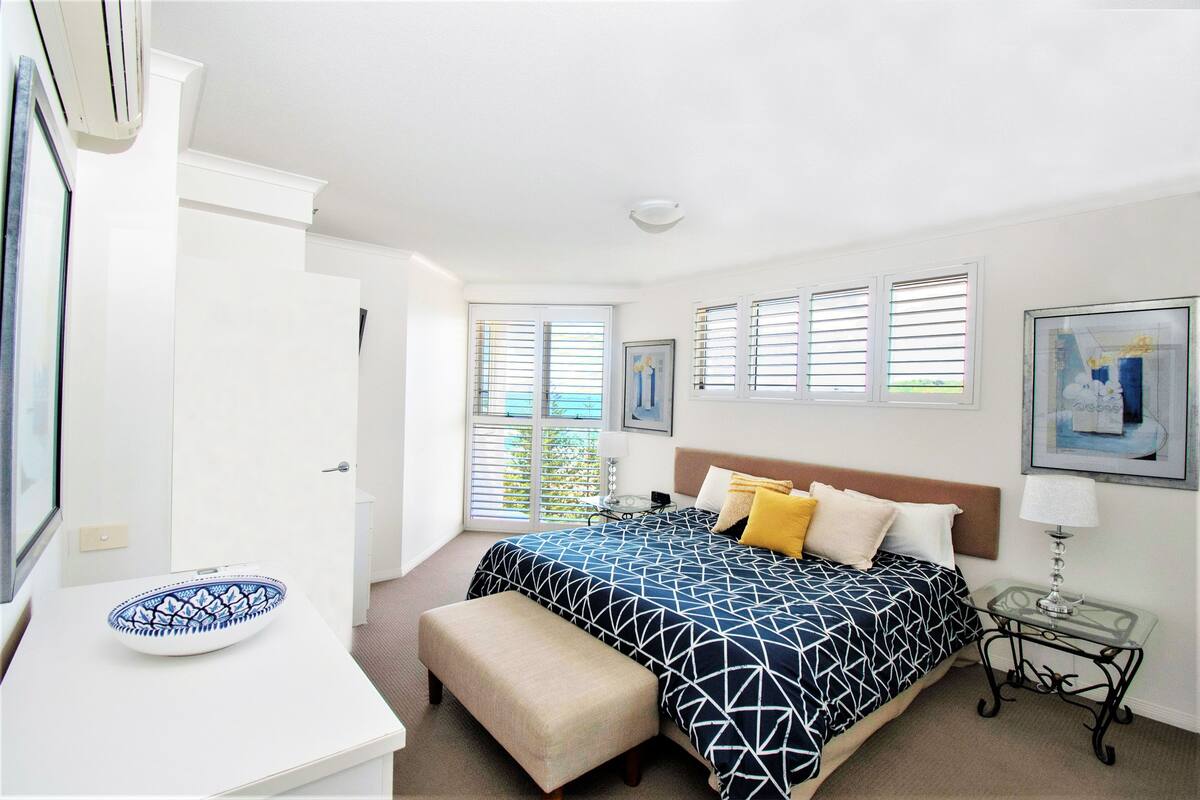 Um quarto com uma cama, mesa de cabeceira, banco e uma tigela sobre a mesa.