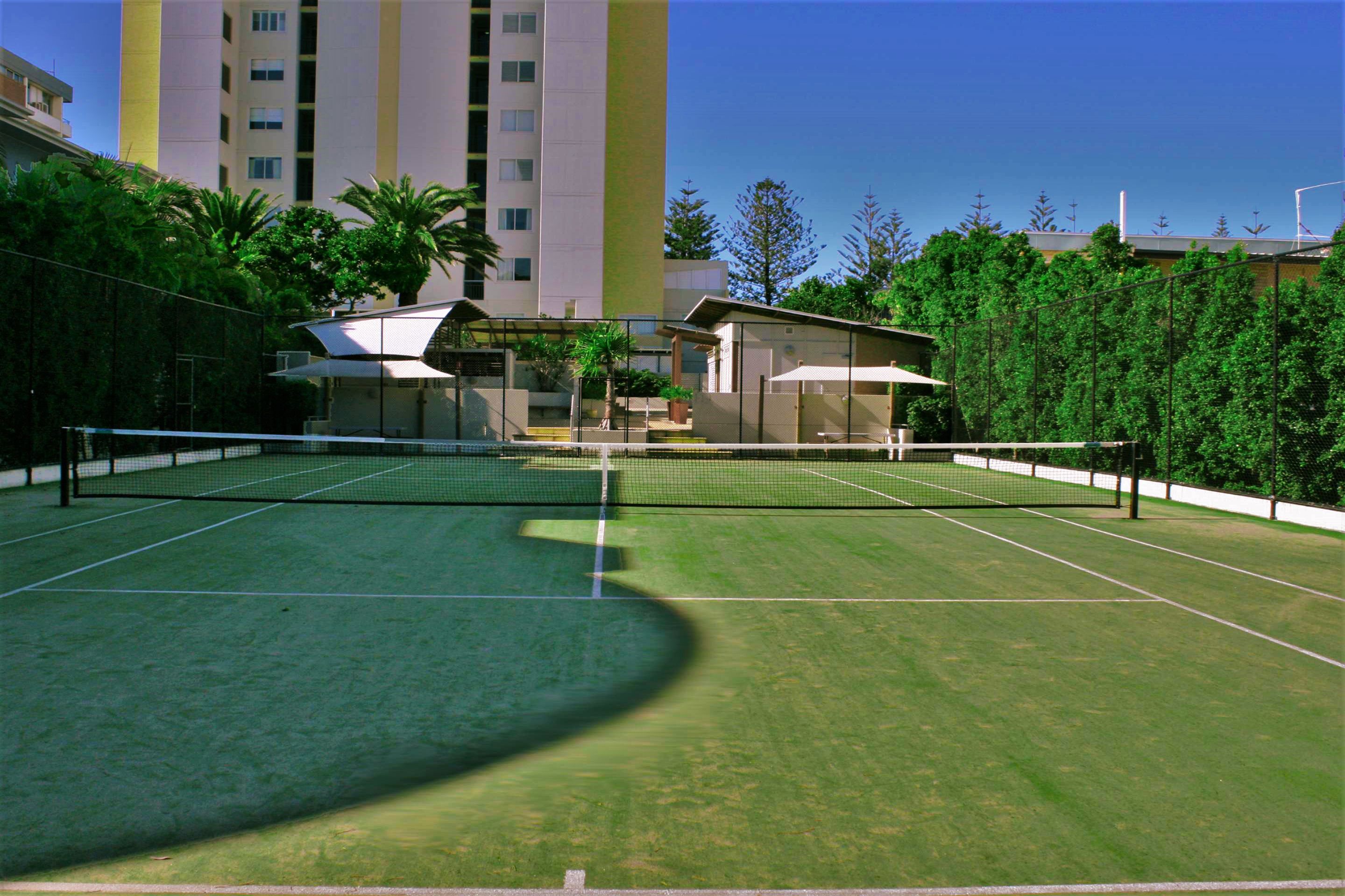 An outdoor tennis court with a net, surrounded by greenery and a building in the background.
