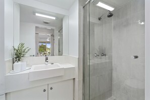 A modern bathroom with a large mirror, a white sink, and a glass-enclosed shower. - The Hill Apartments (Currumbin)