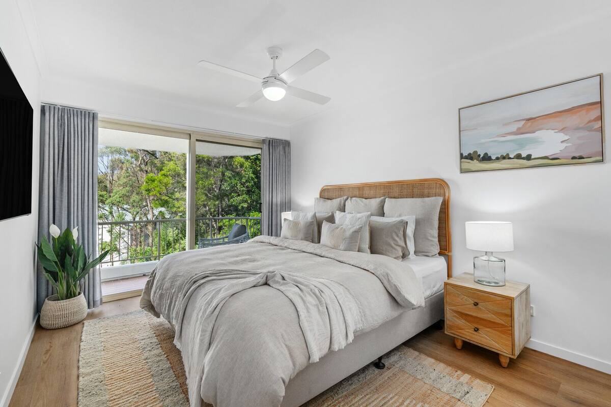 A bedroom with a bed, a nightstand, a ceiling fan, a window with curtains, a plant, and a painting on the wall.