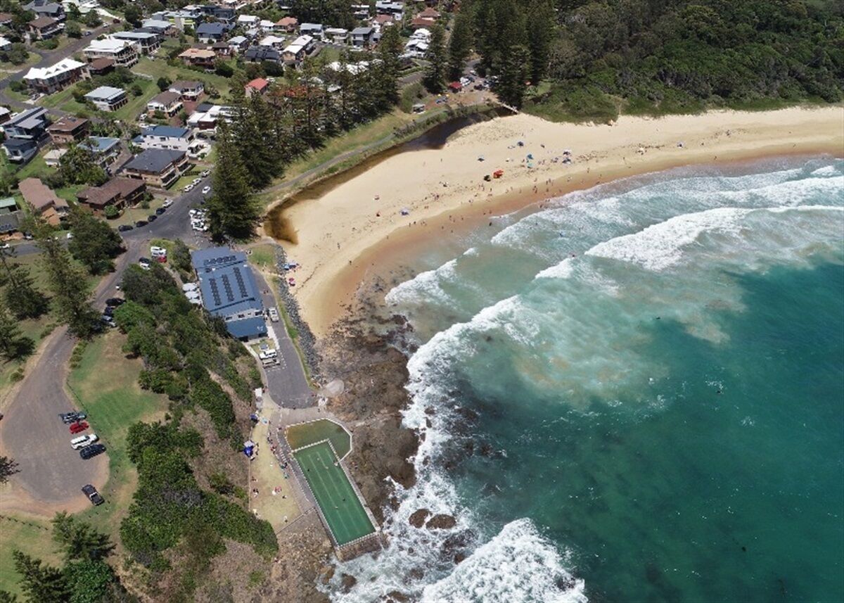An aerial view of a coastal area with a sandy beach, a small parking lot, and a tennis court.