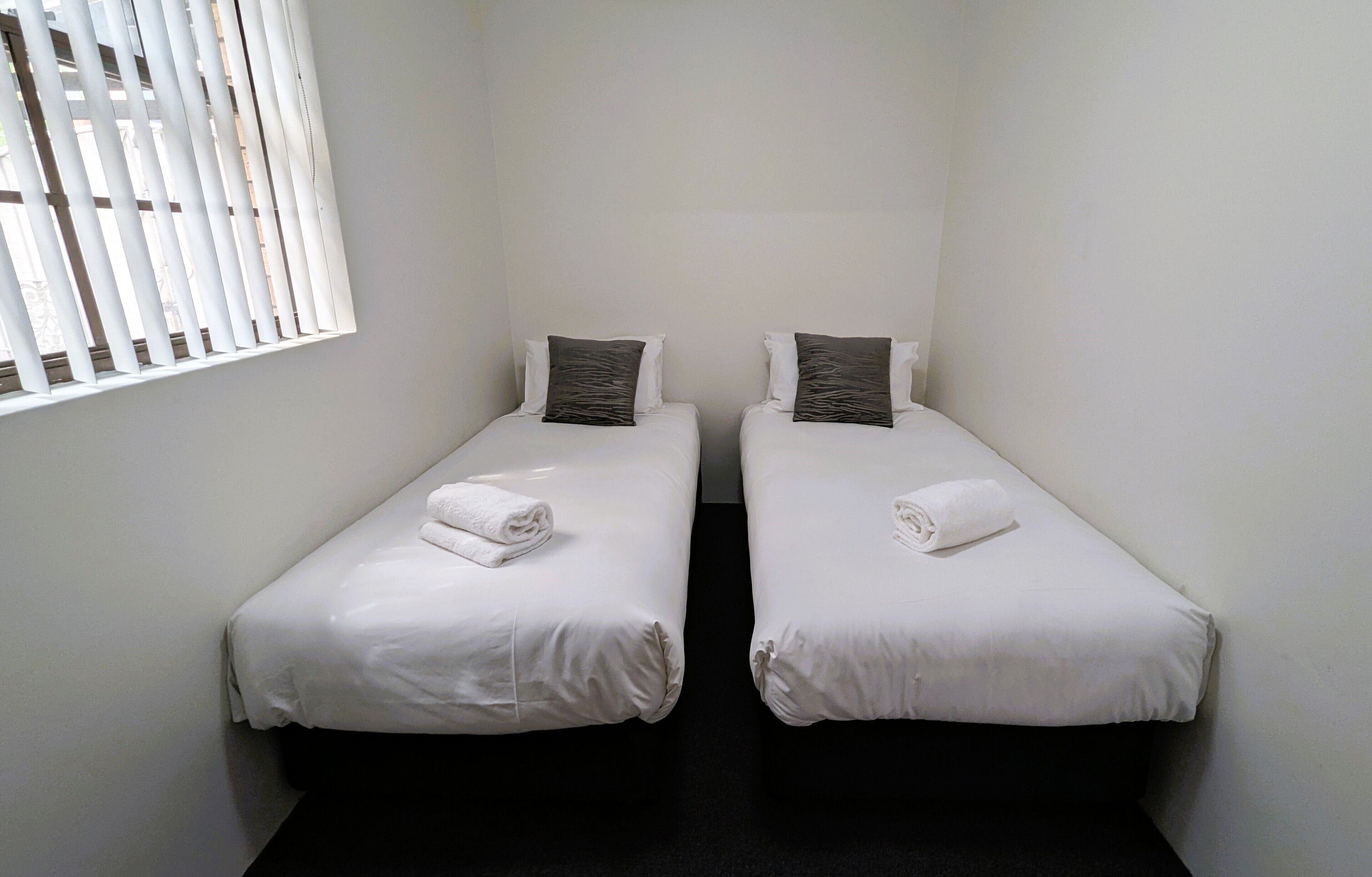 Une petite chambre propre, avec deux lits simples, du linge de lit blanc et une fenêtre avec des stores.
