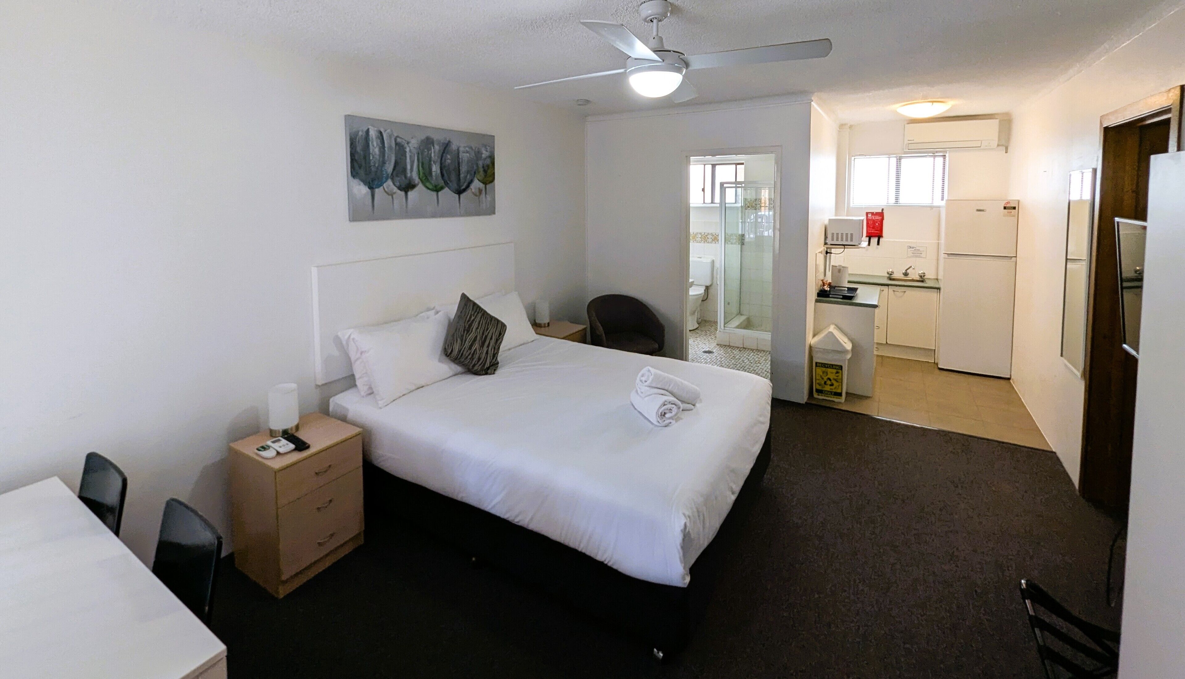 A hotel room with a bed, bedside table, chair, and a painting on the wall. There is a kitchenette in the adjacent room.
