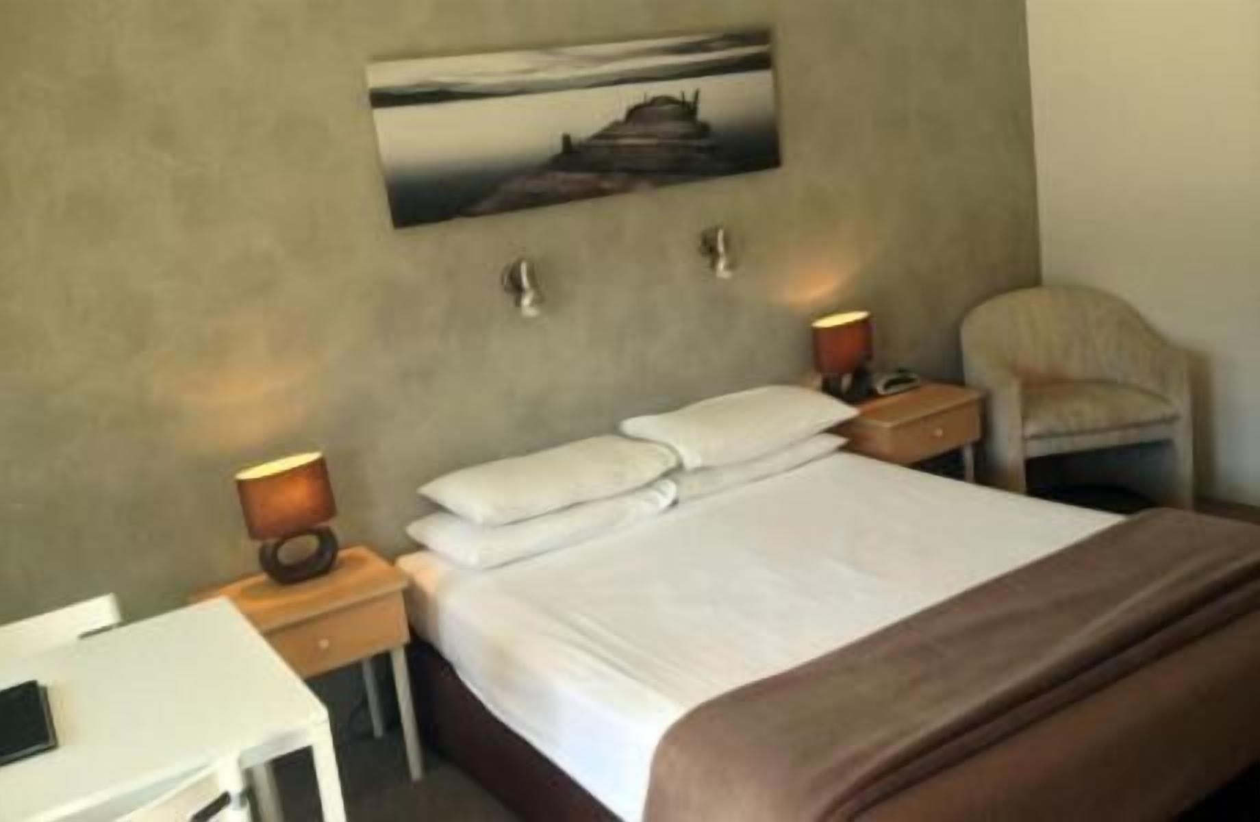 Habitación de hotel con una cama, dos mesitas de noche con lámparas, una silla y un cuadro en la pared.