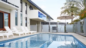 A pool area with white lounge chairs, a sign for Bay Beach Motel, and a palm tree. - Bay Beach Motel (Byron Bay)