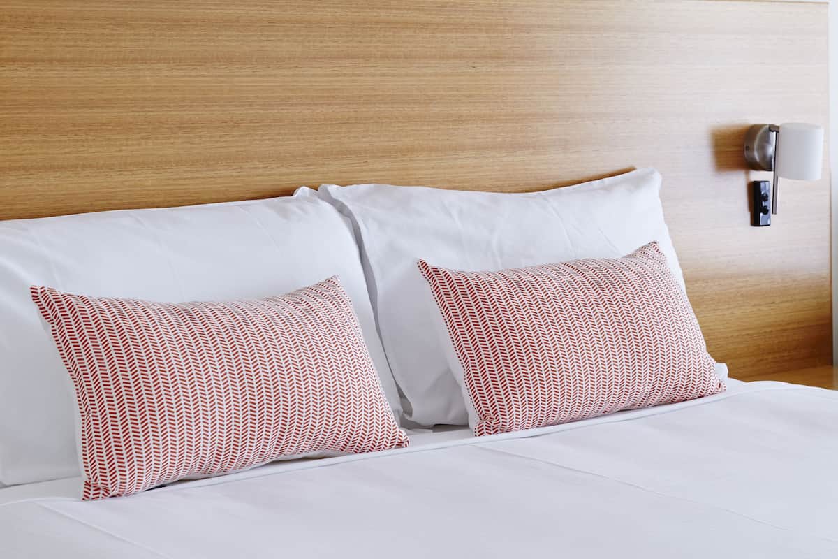 Una cama bien hecha con sábanas blancas y almohadas estampadas, un cabecero de madera y una lámpara fijada a la pared.