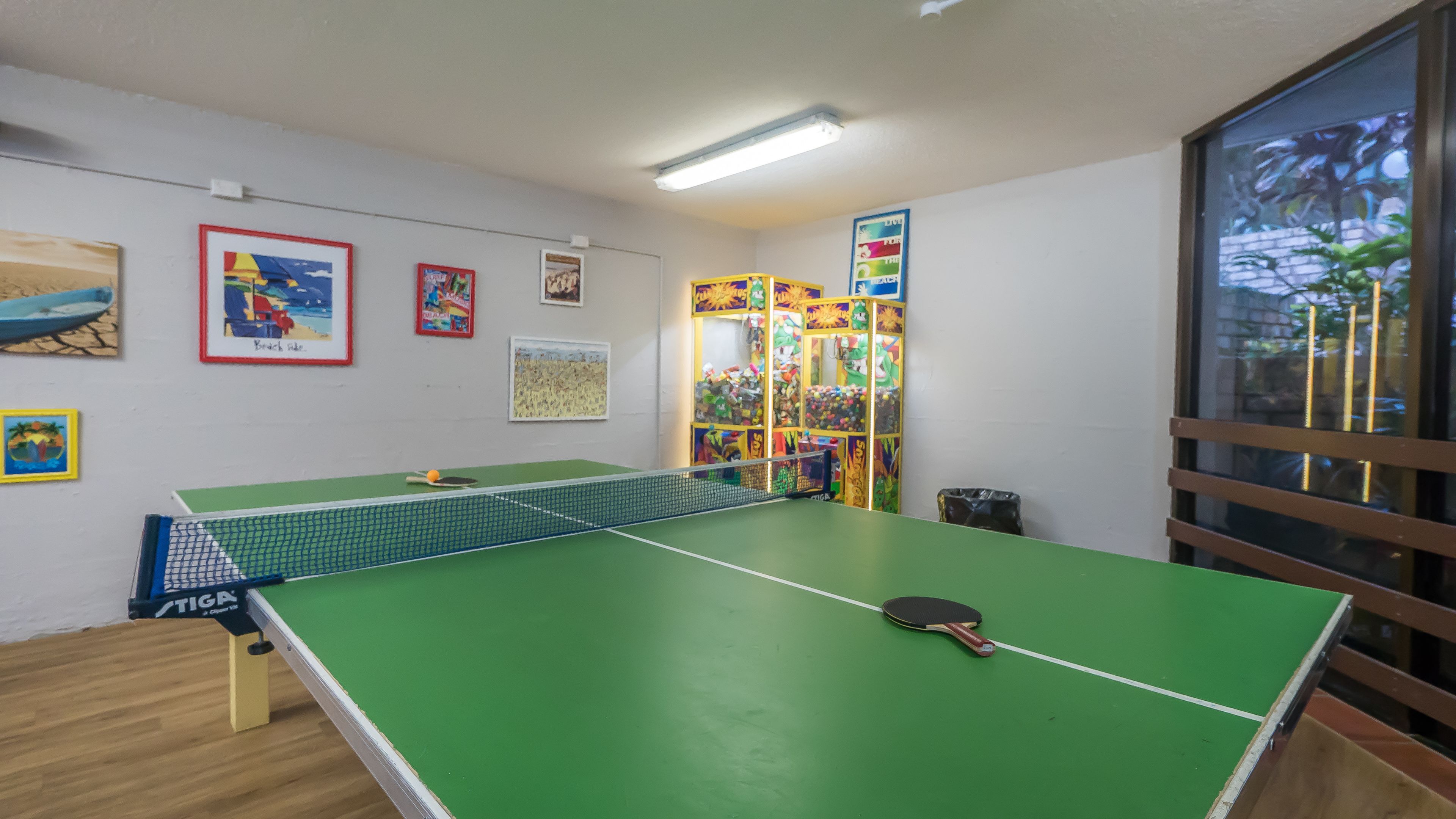 Ein Tischtennis-Tisch mit Netz, umgeben von gerahmten Kunstwerken an den Wänden, und ein Fenster mit Jalousien.