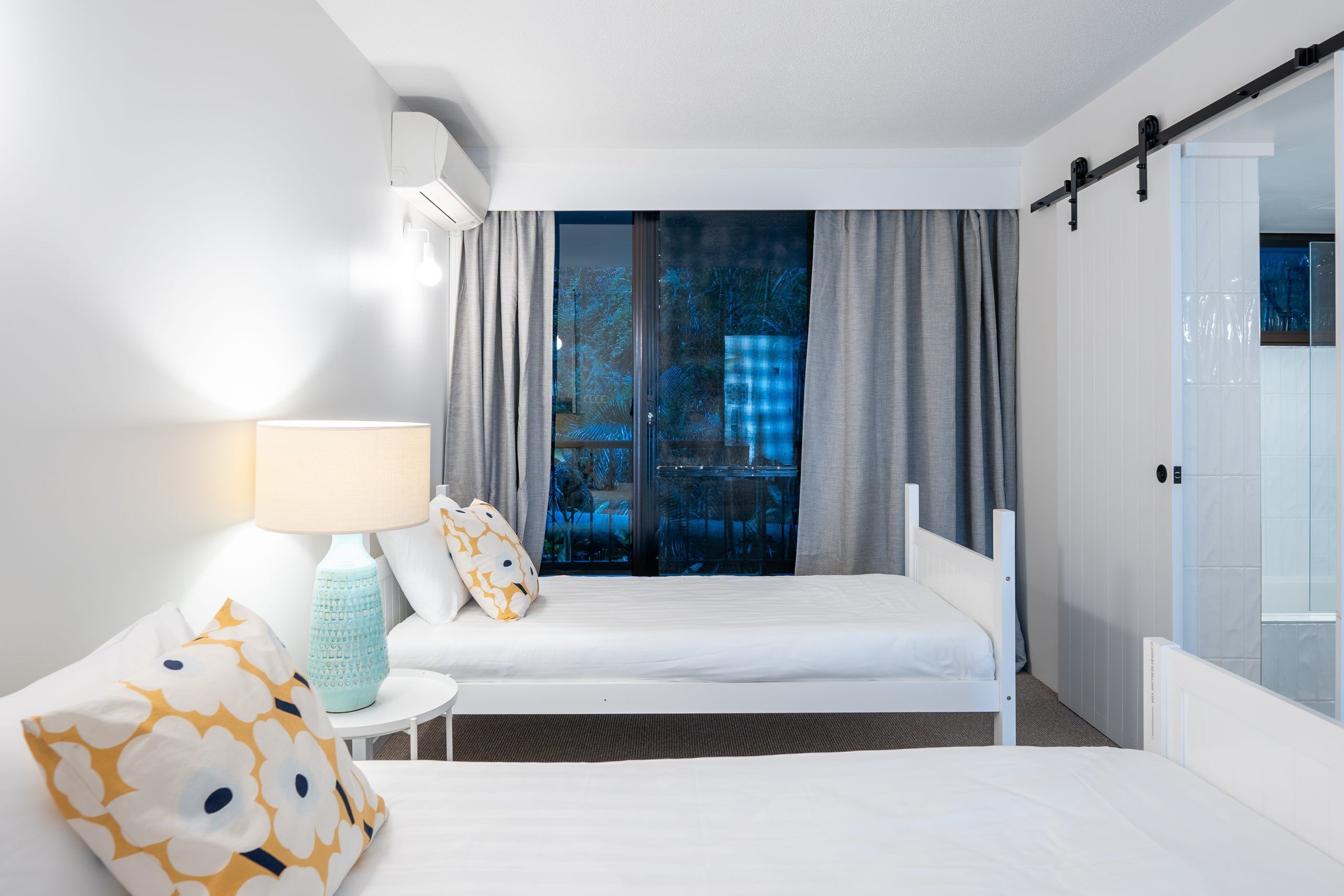 A hotel room with two beds, a small table with a lamp, and a view of a balcony with a checkered tablecloth.