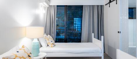 A hotel room with two beds, a small table with a lamp, and a view of a balcony with a checkered tablecloth.
