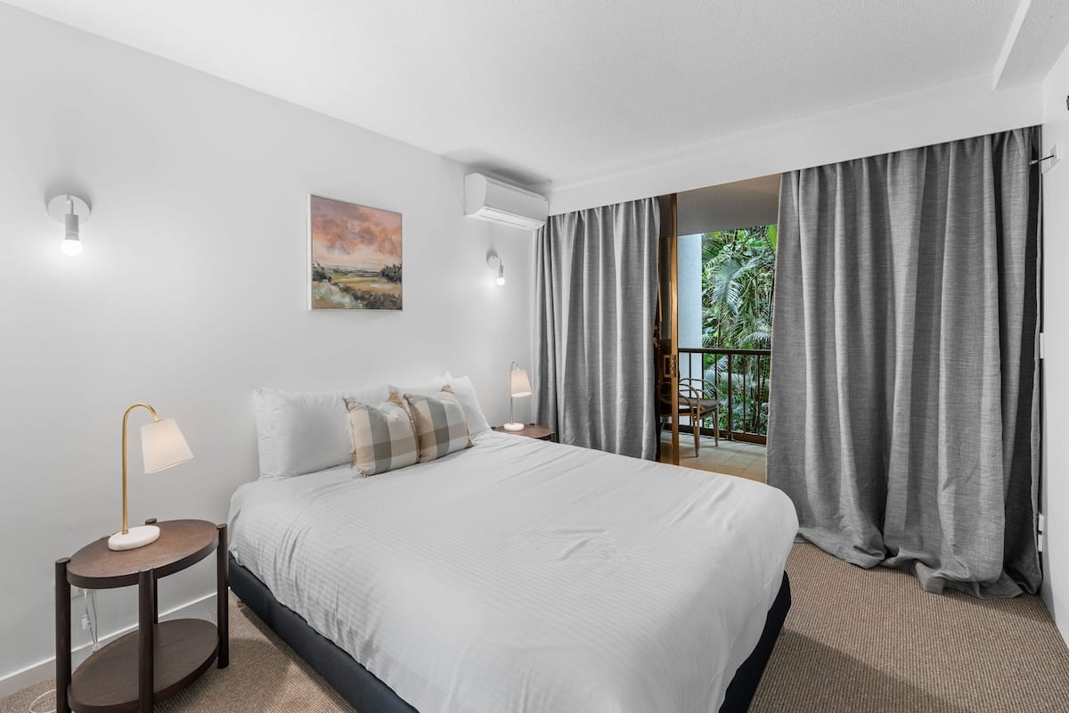 Une chambre d’hôtel avec un grand lit, deux tables de chevet, un tableau au mur et une vue sur la verdure par la fenêtre.