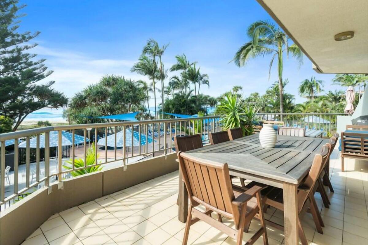 Une table et des chaises en bois sur un balcon donnant sur une piscine et des palmiers.