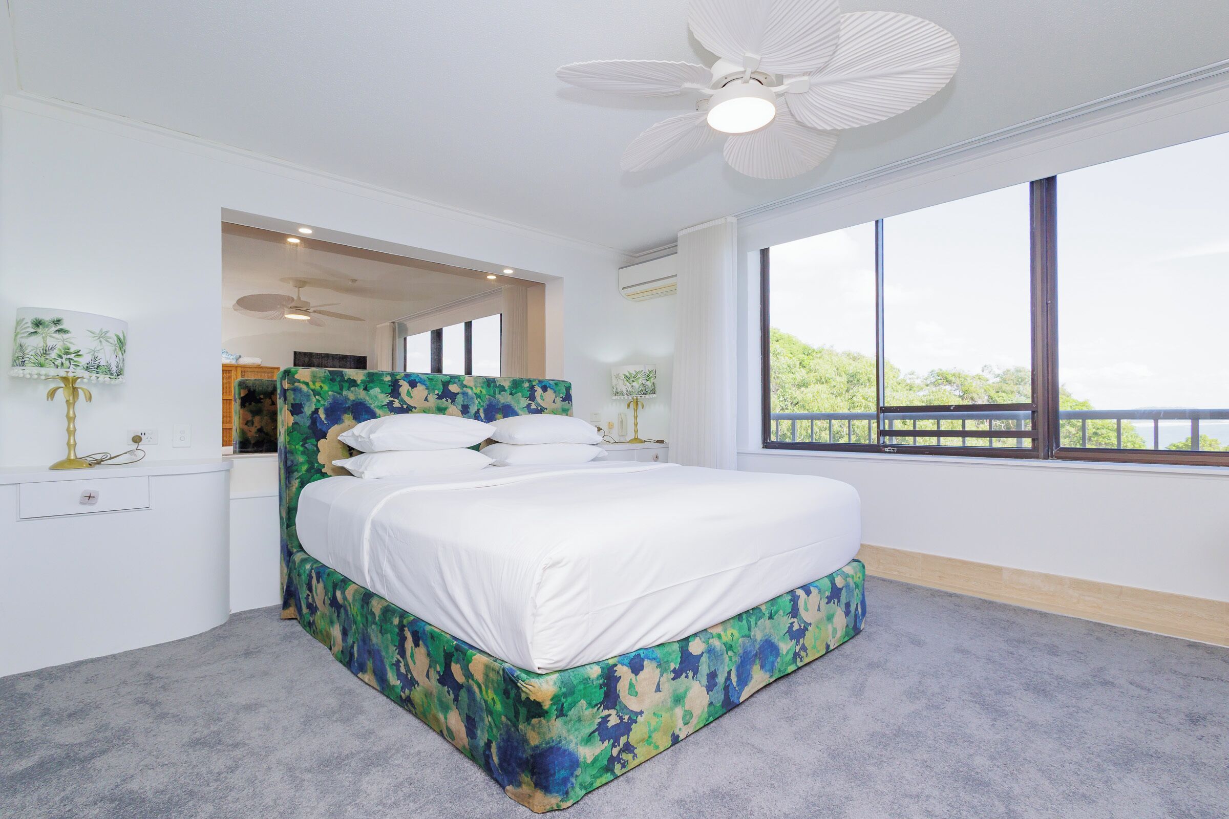 Une chambre moderne avec un grand lit, un ventilateur de plafond et une vue sur la verdure par la fenêtre.