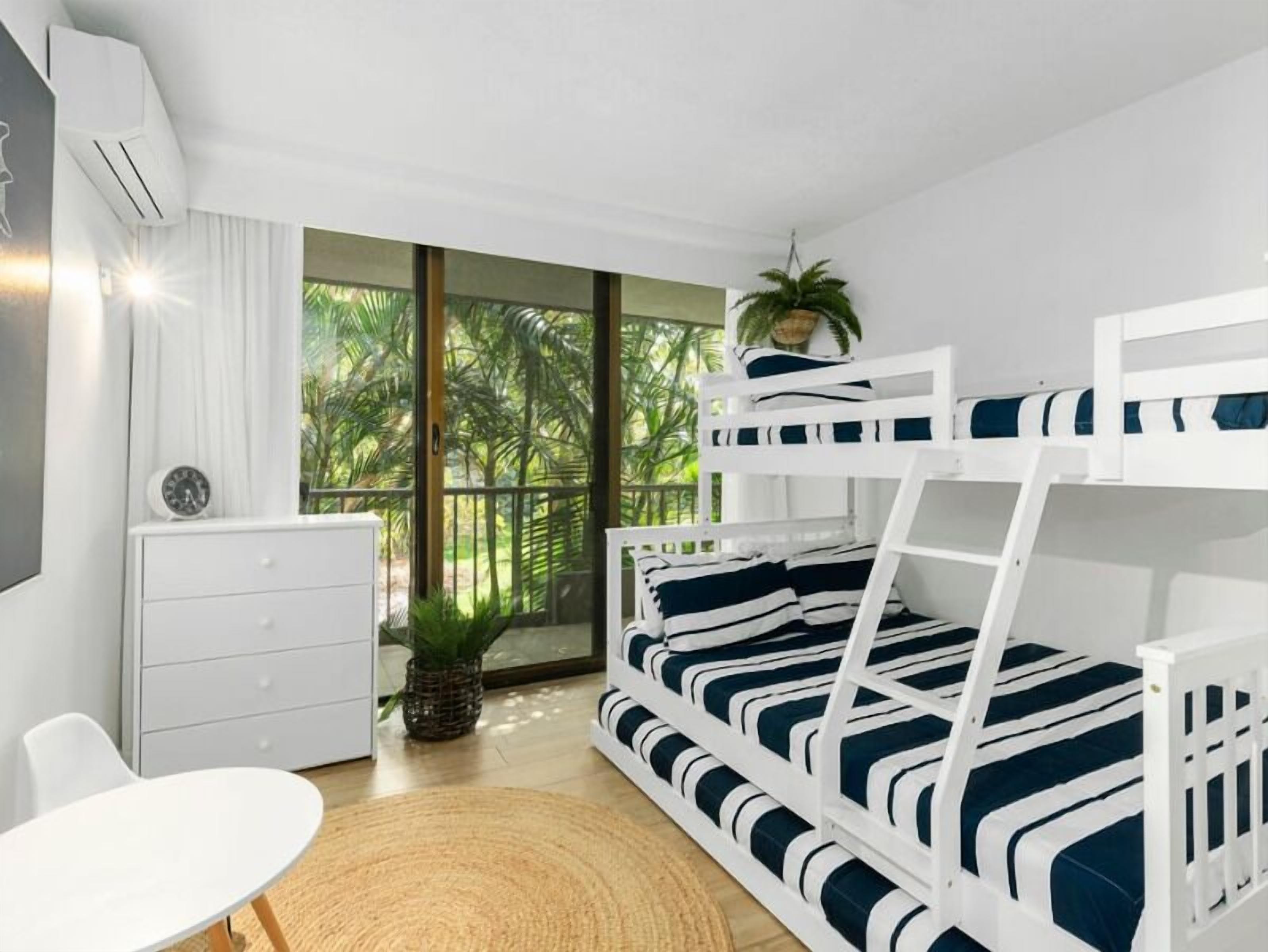 Ein modernes Schlafzimmer mit Etagenbett, kleinem Schreibtisch, Stuhl und großem Fenster mit Blick auf Grünflächen.