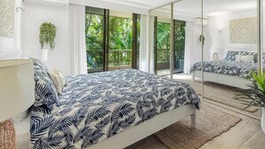 Un dormitorio con cama, un espejo grande y una puerta corrediza de vidrio que da a un balcón con vegetación.