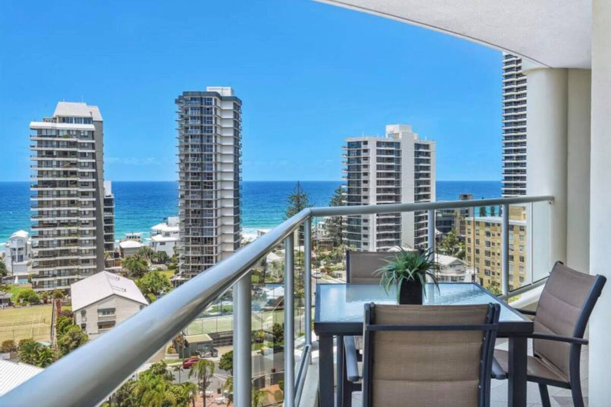 A balcony with a table and chairs, overlooking a coastal cityscape with tall buildings and the ocean.
