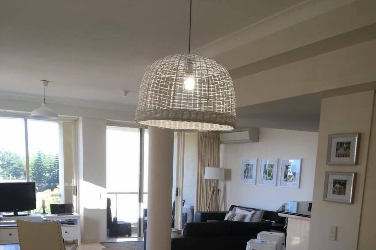 A modern dining area with a wooden table and white chairs, a hanging woven light fixture, and a view of the living room in the background.