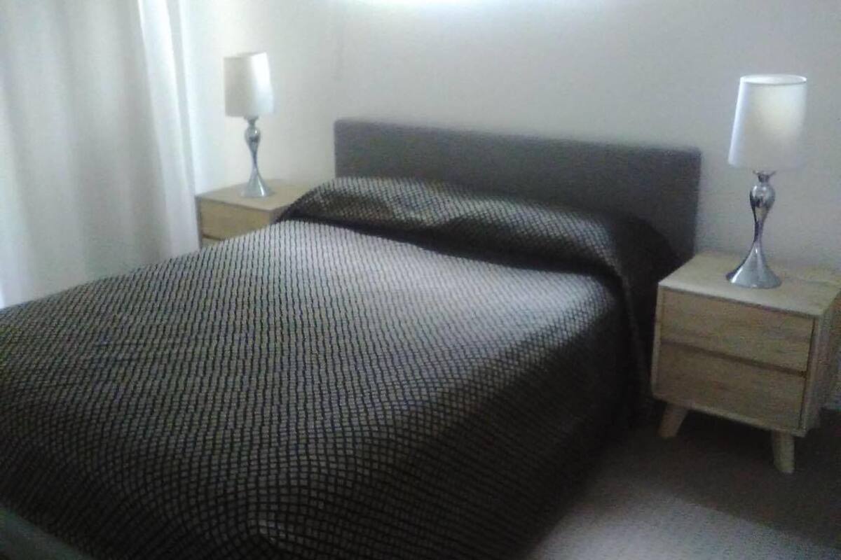 Una cama bien tendida con una colcha oscura estampada, una mesita de noche con lámpara y una ventana con cortinas.