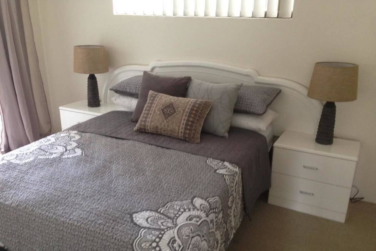 Una cama bien tendida con una colcha estampada, dos lámparas de noche y una mesita de noche con cajones.