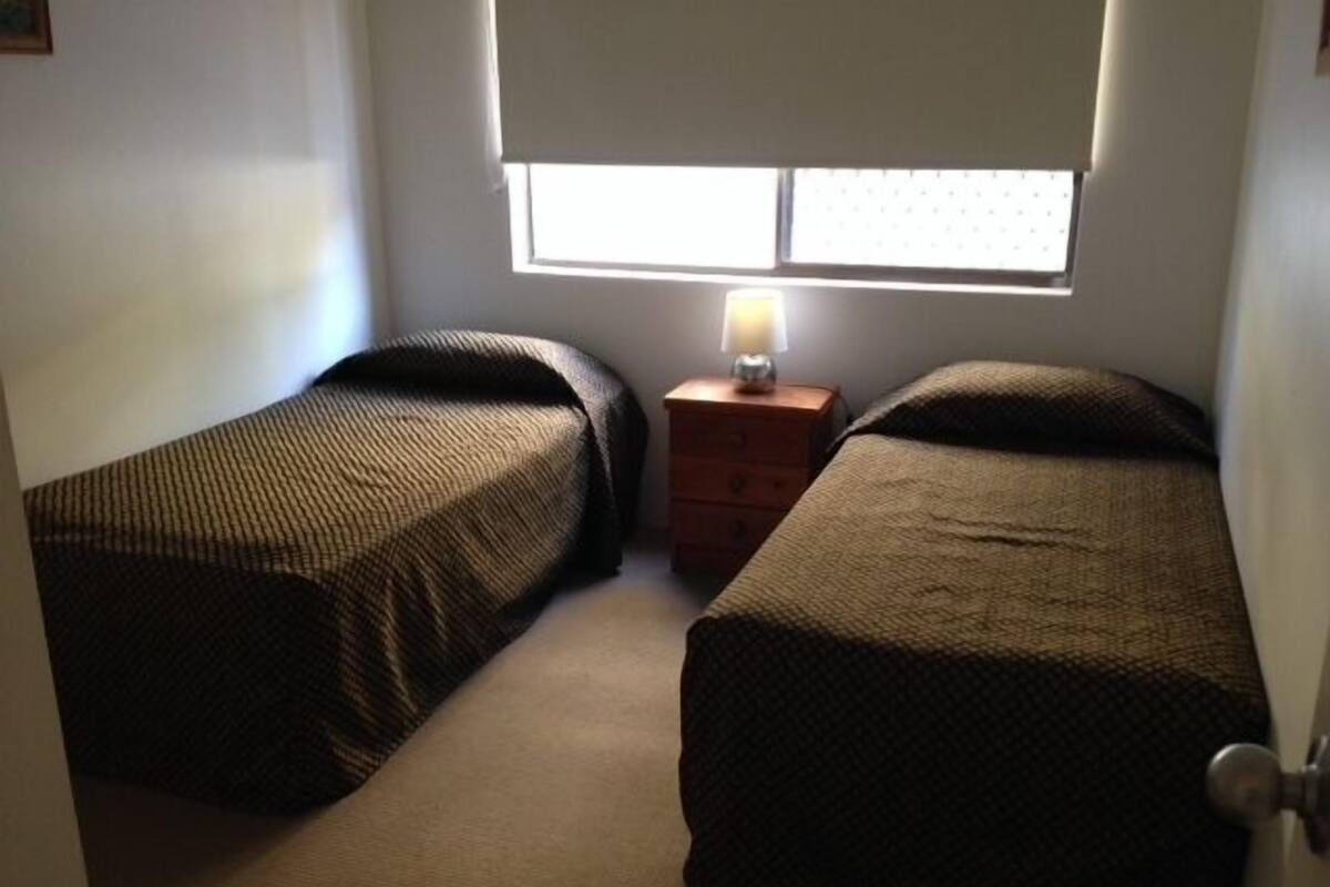 Una habitación pequeña con dos camas individuales, una mesita de noche con lámpara y una ventana con persianas.