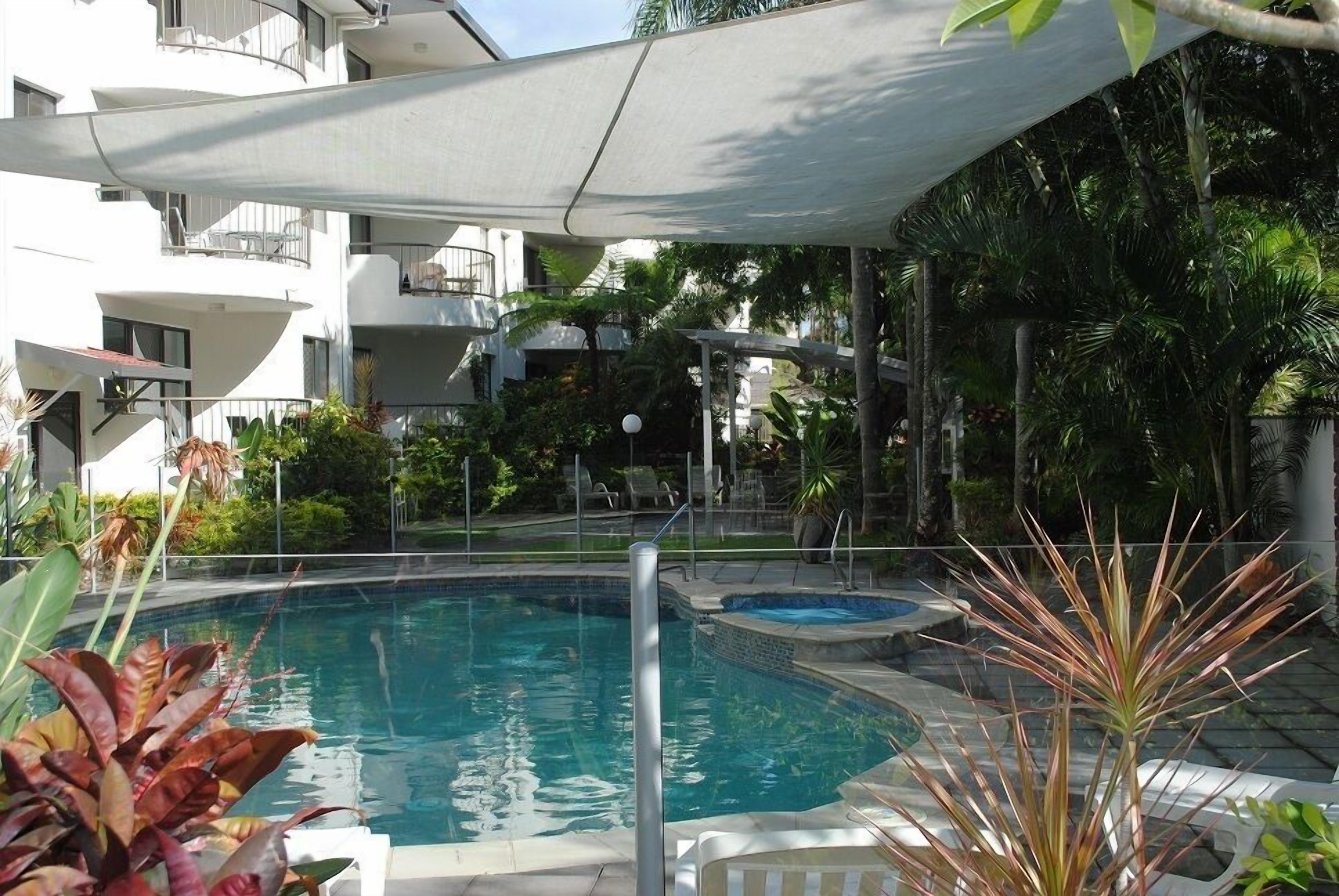 Una piscina con sombrilla, rodeada de vegetación exuberante y un edificio de varios pisos al fondo.