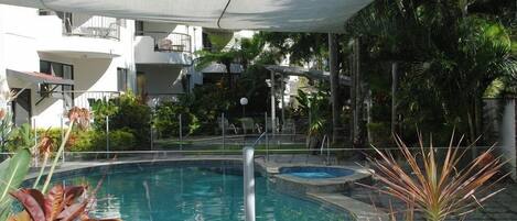 Una piscina con sombrilla, rodeada de vegetación exuberante y un edificio de varios pisos al fondo.