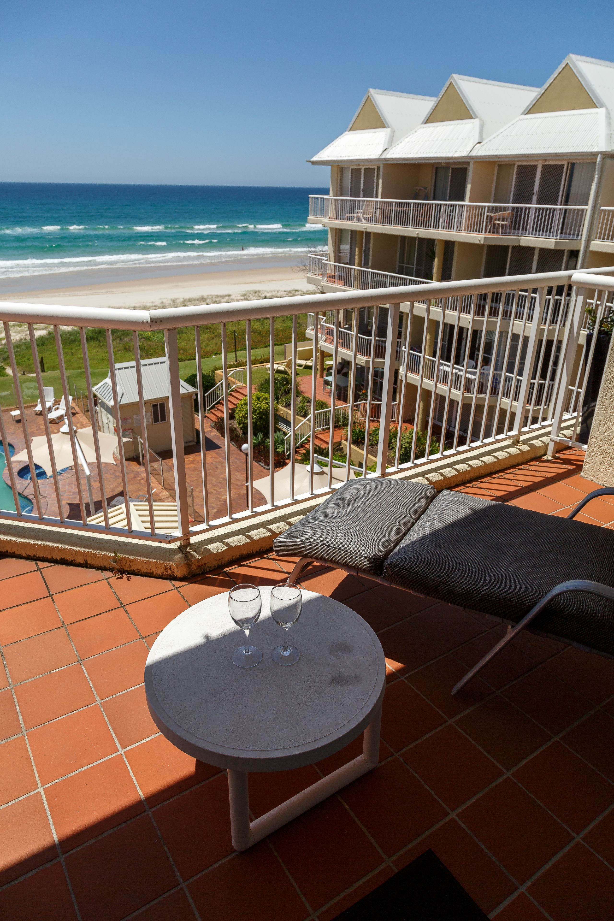 A balcony with a table and chairs, overlooking a beach and a building with multiple balconies.